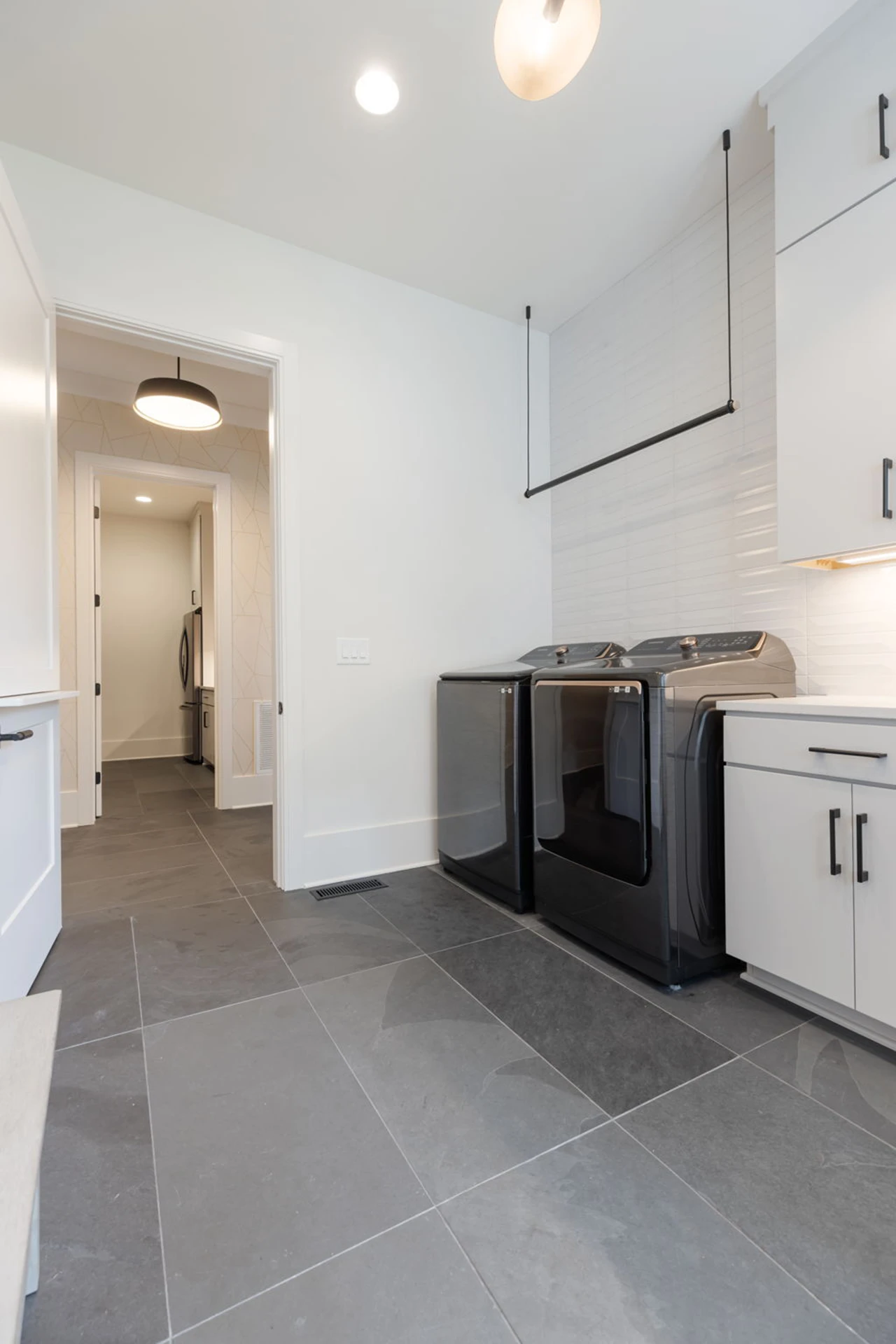 Laundry room with white cabinets, black washer and dryer, gray tile floor, and a hallway to a bathroom