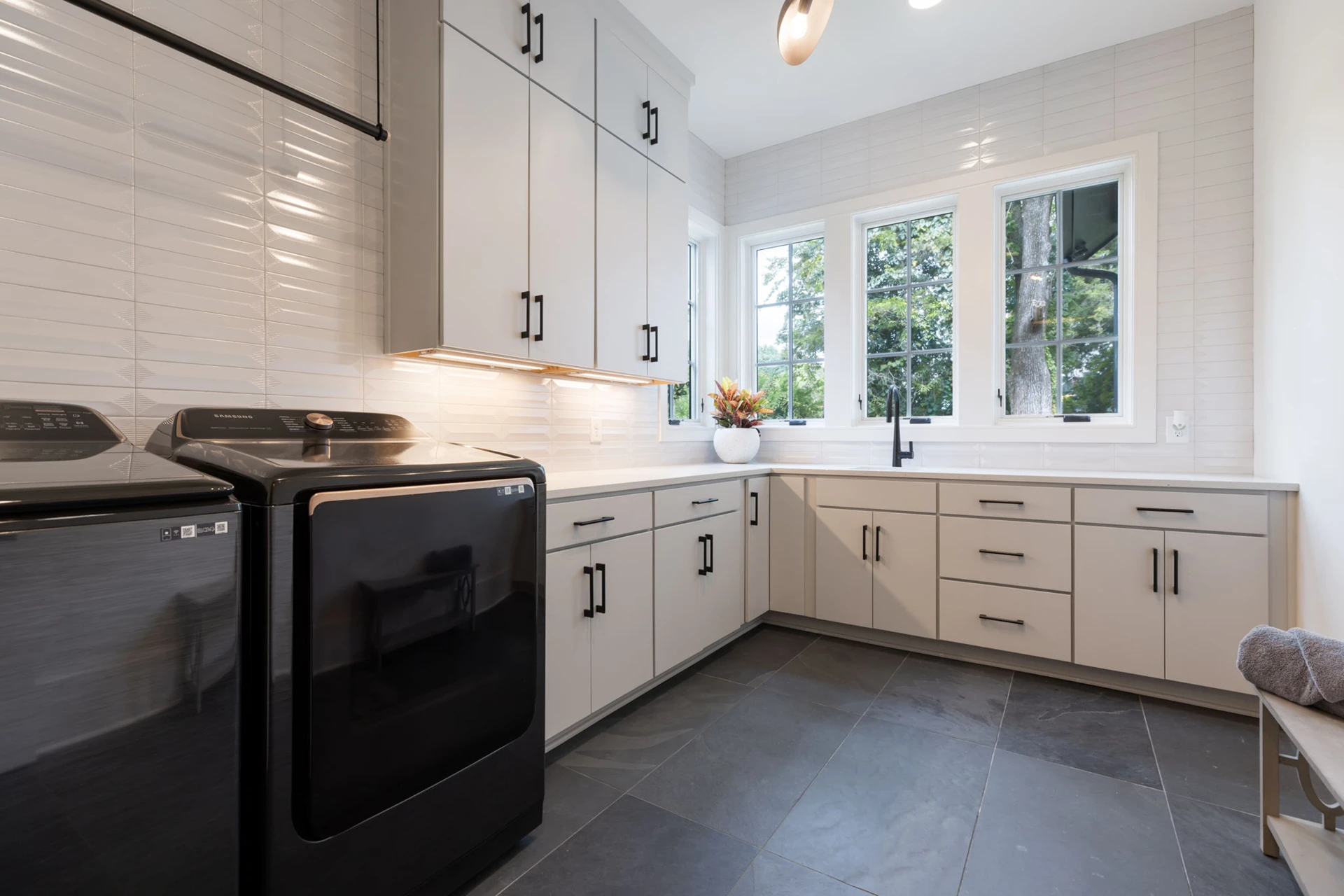 Laundry room with light gray cabinets, black washer and dryer, white tile backsplash, and a window
