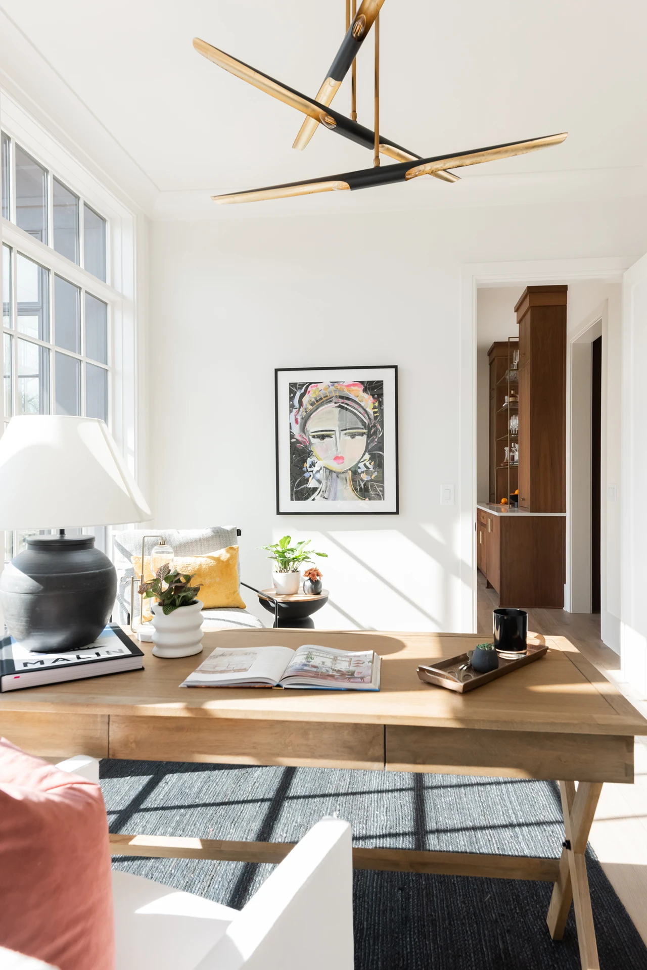 Home office corner with a wood X-leg desk, black-and-gold linear chandelier, and framed portrait art