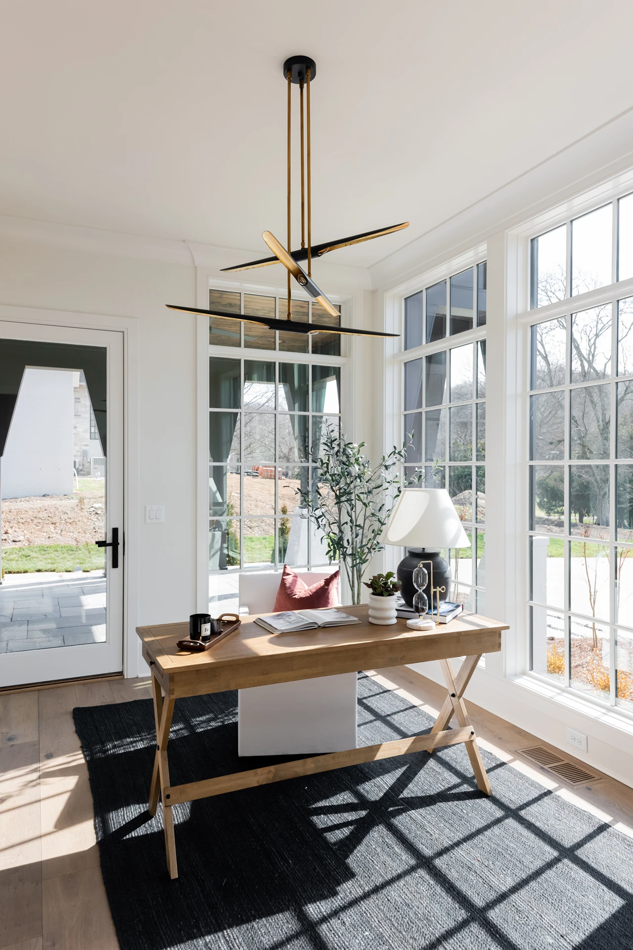 Home office with wood X-leg desk, black-and-gold sputnik chandelier, wall of windows, and a striped rug