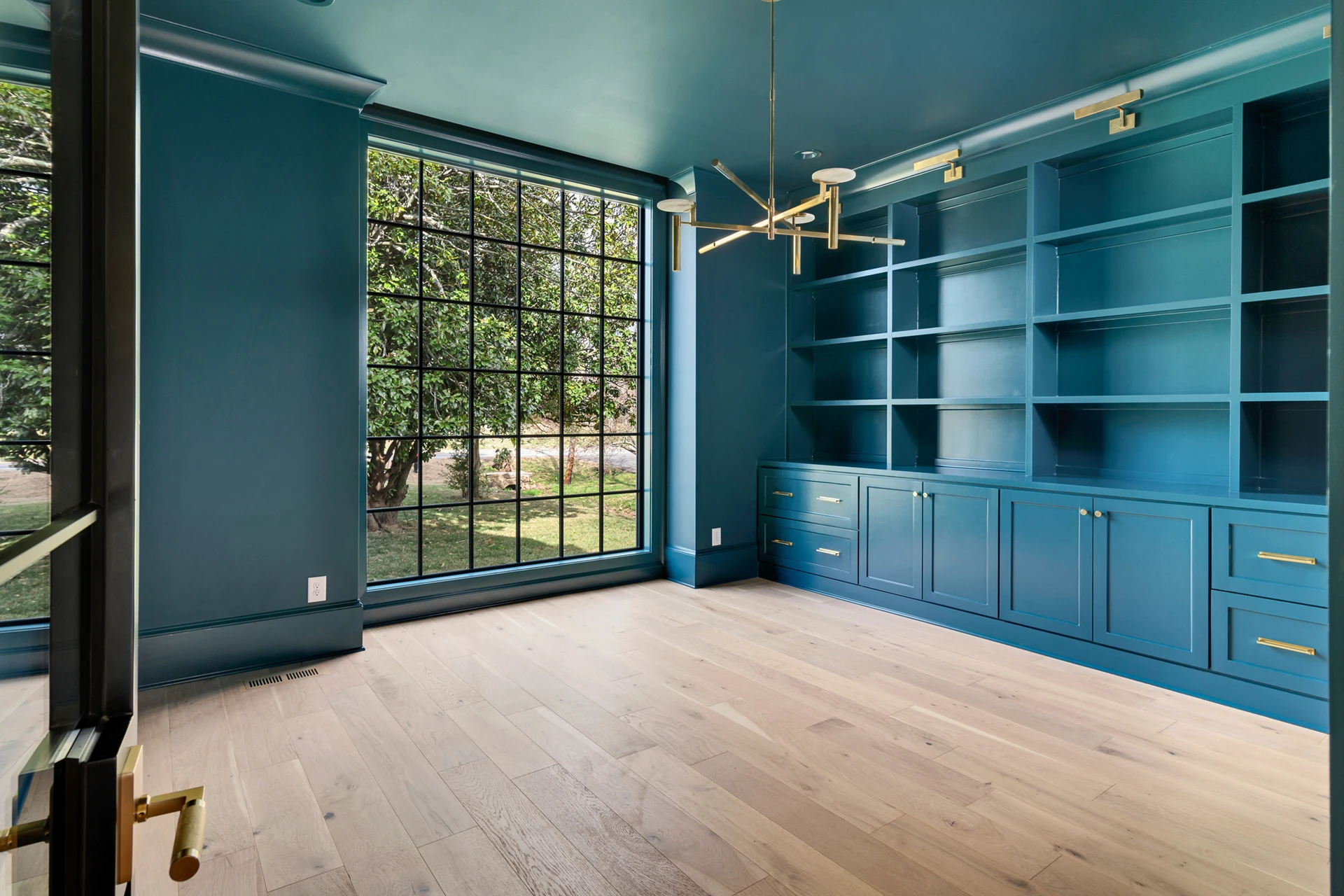Empty study with teal walls and ceiling, built-in teal shelving, large black-grid window, and sputnik chandelier
