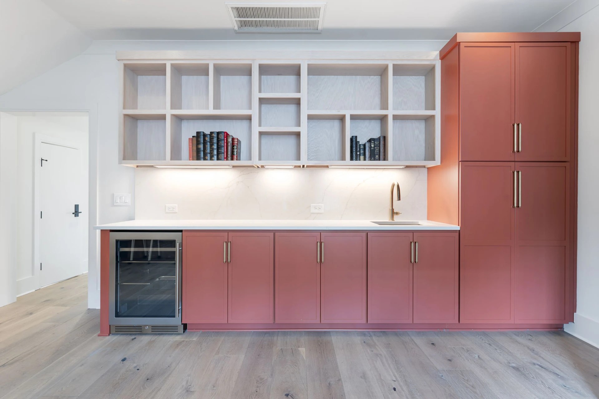 Bonus room bar with terracotta cabinets, open shelving of books, beverage fridge, and a gold faucet