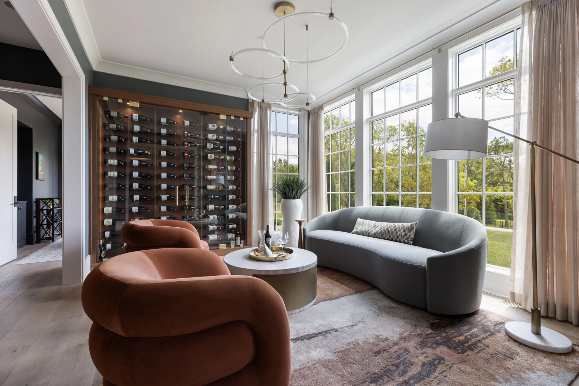 Sitting room with wine rack wall, curved gray sofa, rust velvet chair, and a wall of windows