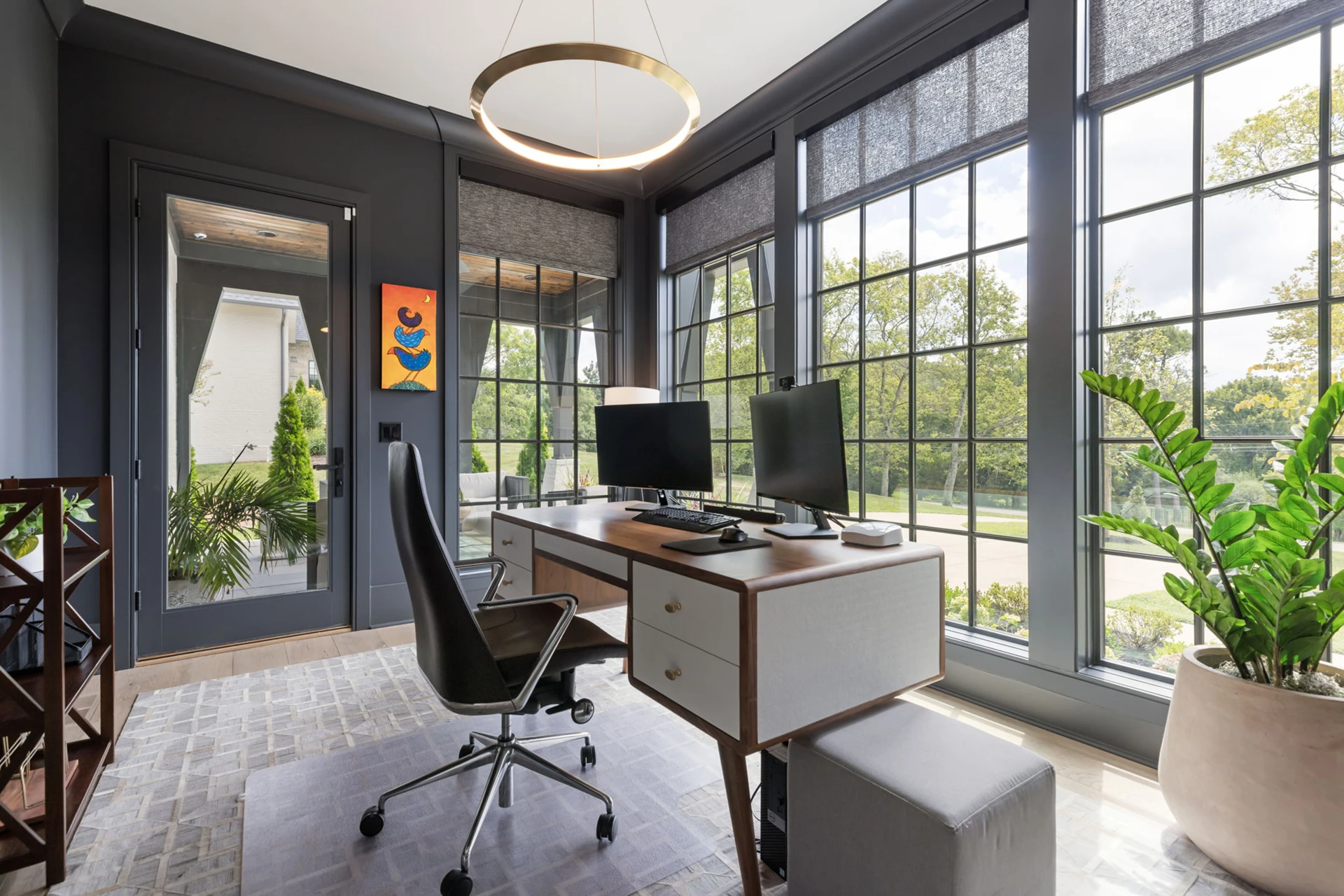 Home office with dark gray walls, gold ring pendant, wood desk with dual monitors, and a wall of windows