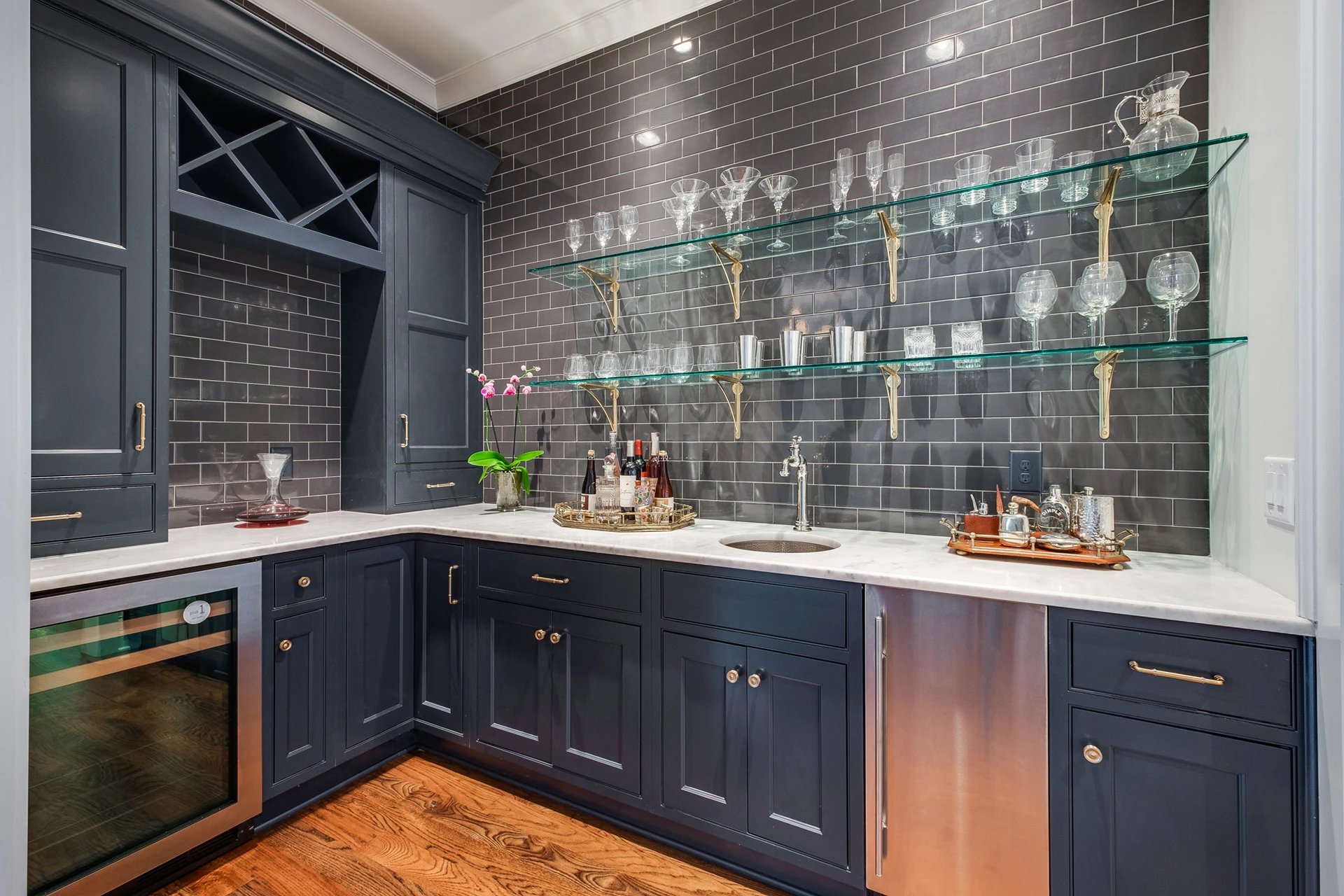 Butler's pantry with navy cabinets, dark subway tile, glass shelves of glassware, and a beverage fridge