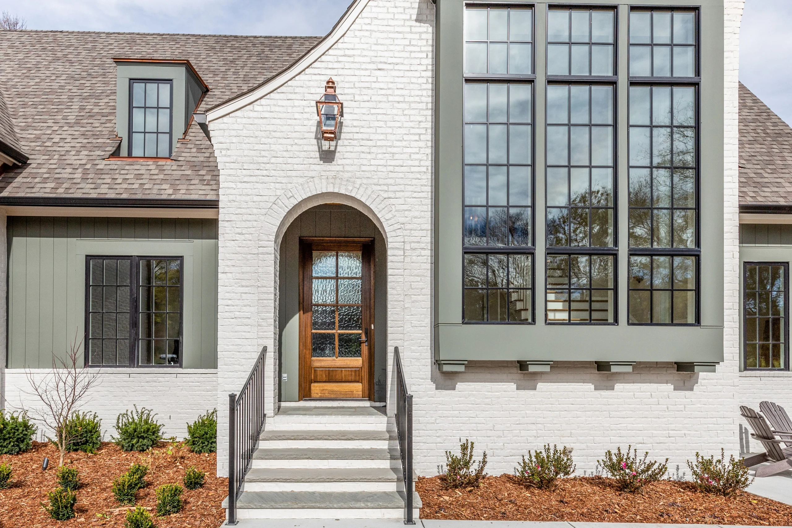 White painted brick home exterior with arched wood front door, black-framed windows, and Adirondack chairs