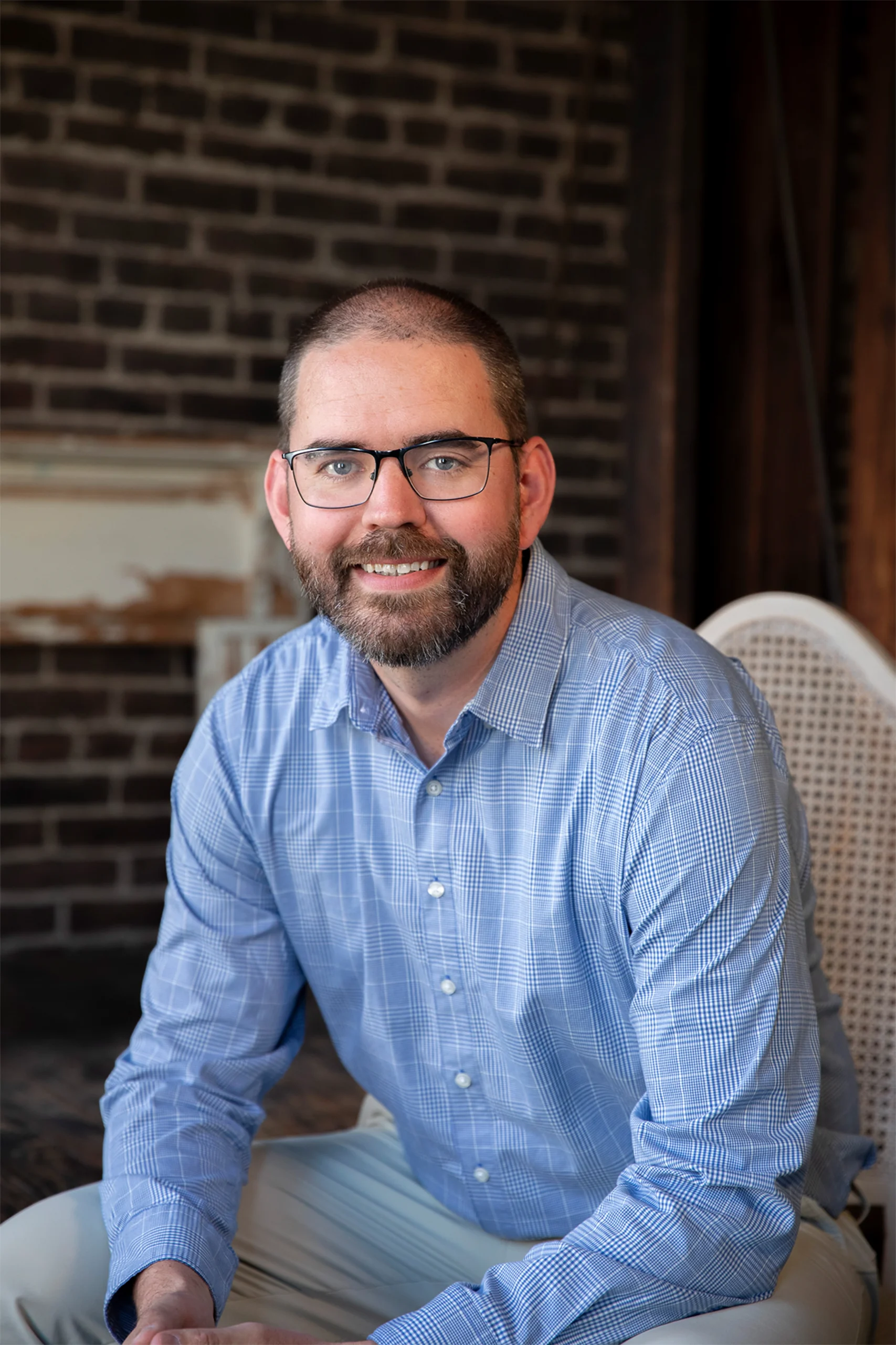 Professional headshot of a smiling man with glasses and beard in a blue shirt against a brick wall