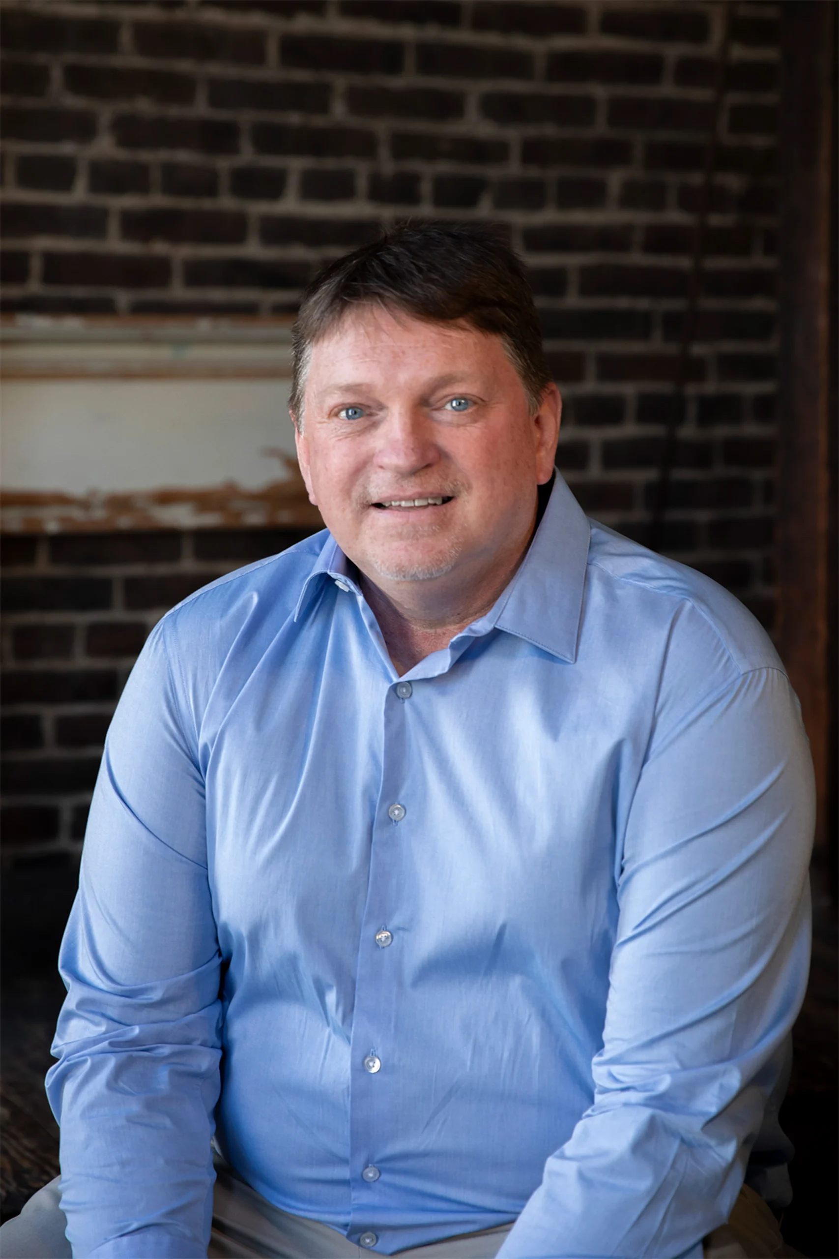 Professional headshot of a smiling man in a light blue shirt against an exposed brick wall