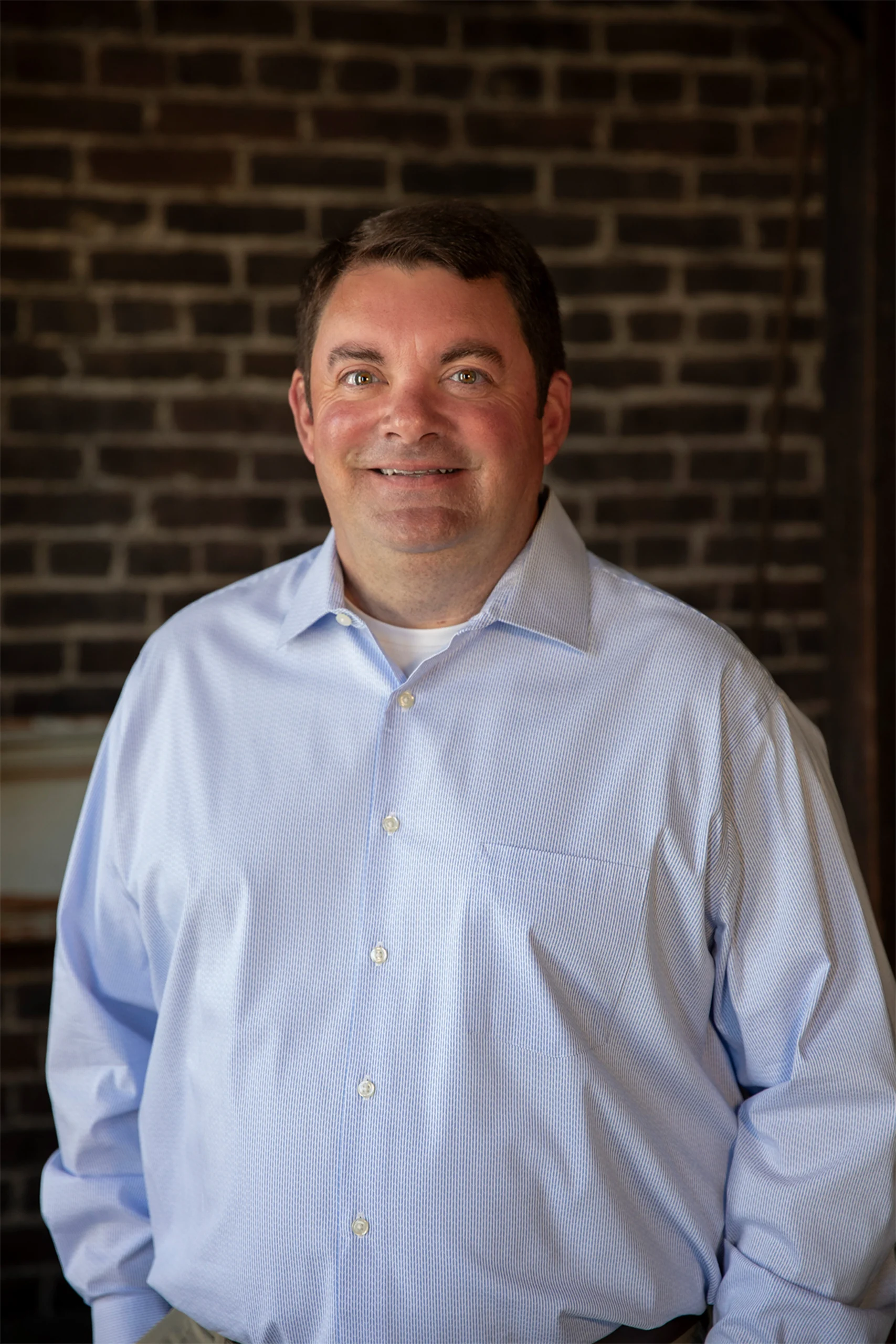 Professional headshot of a smiling man in a light blue striped shirt against an exposed brick wall