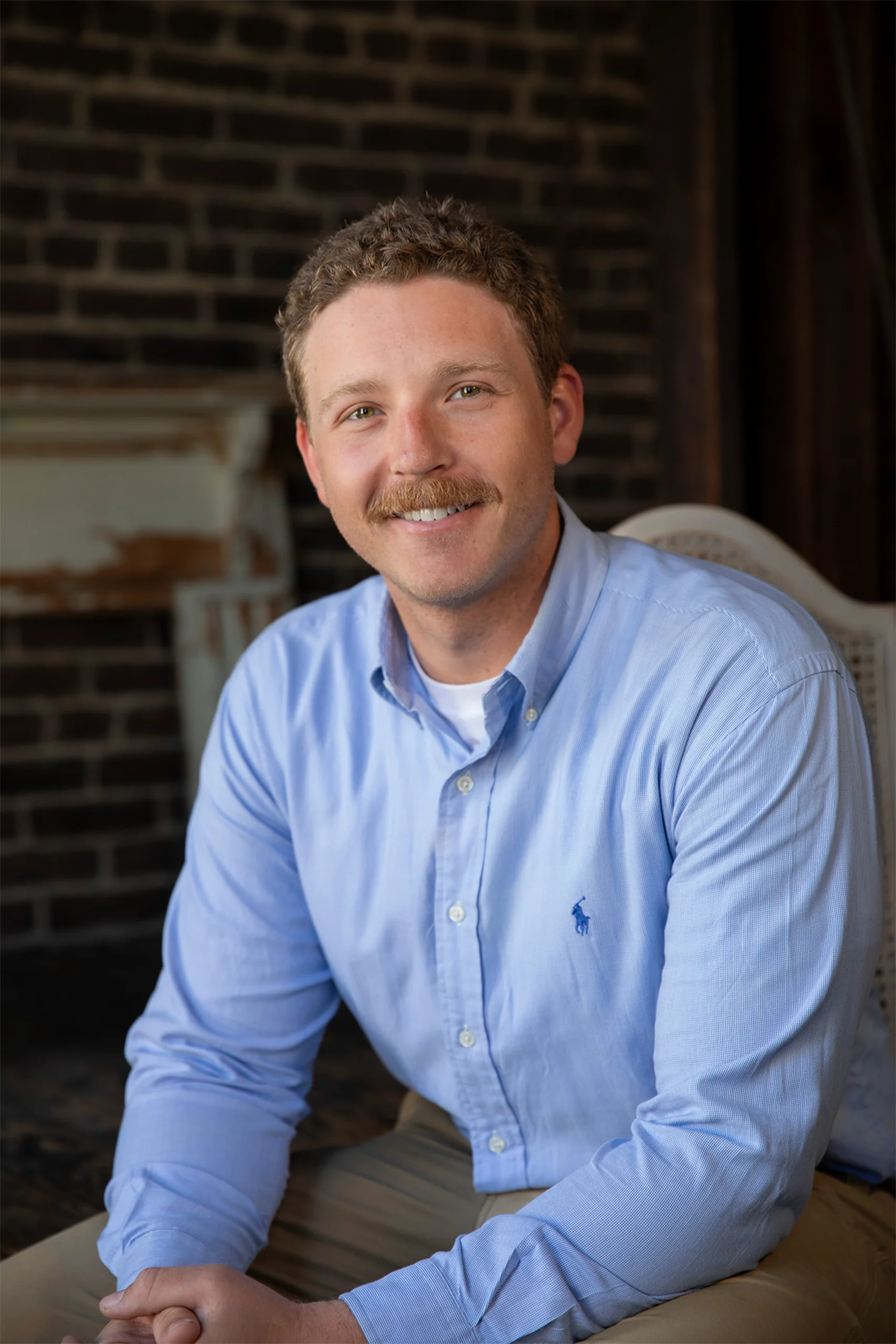 Professional headshot of a smiling man with a mustache in a light blue shirt against a brick wall