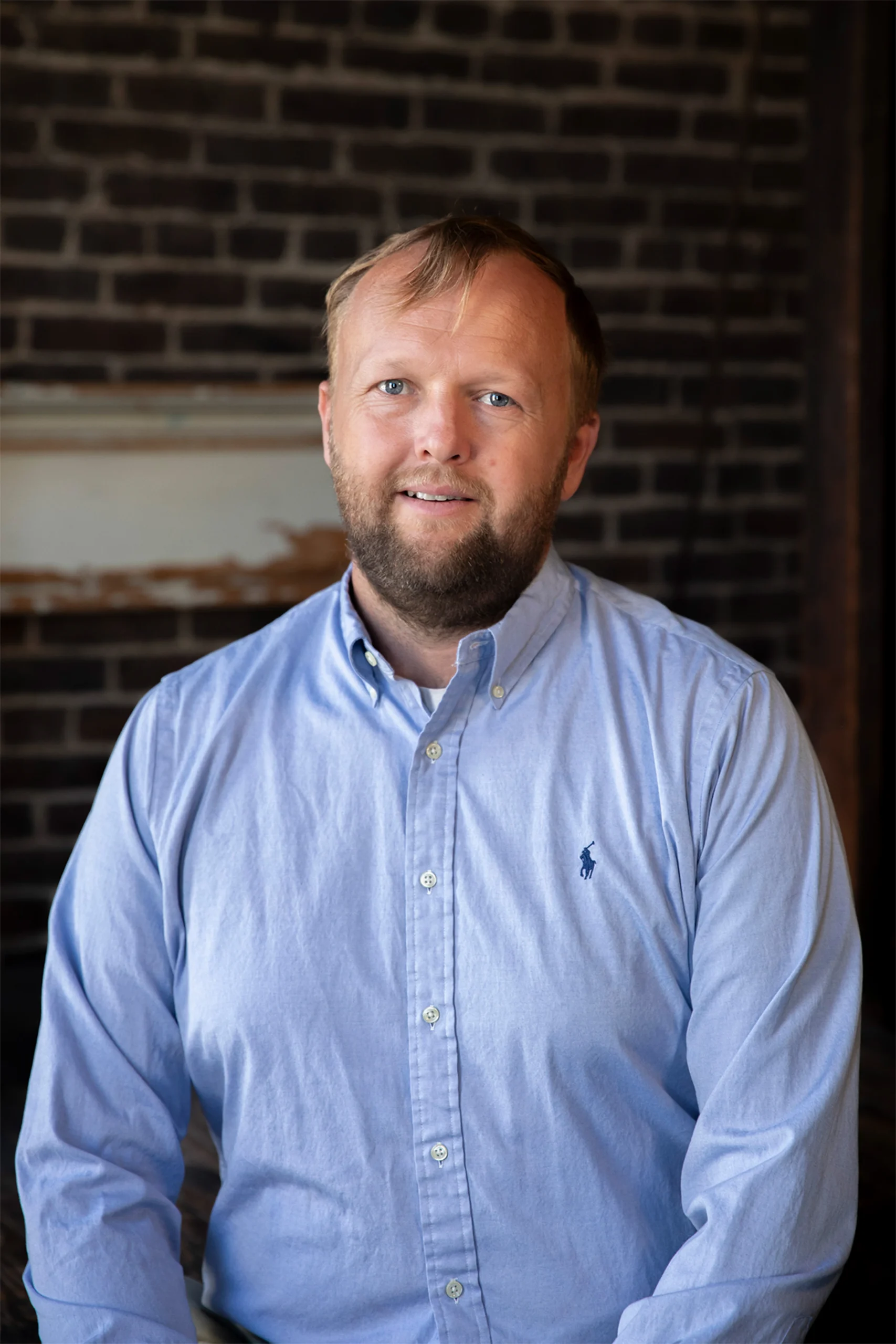 Professional headshot of a smiling bearded man in a light blue shirt against an exposed brick wall