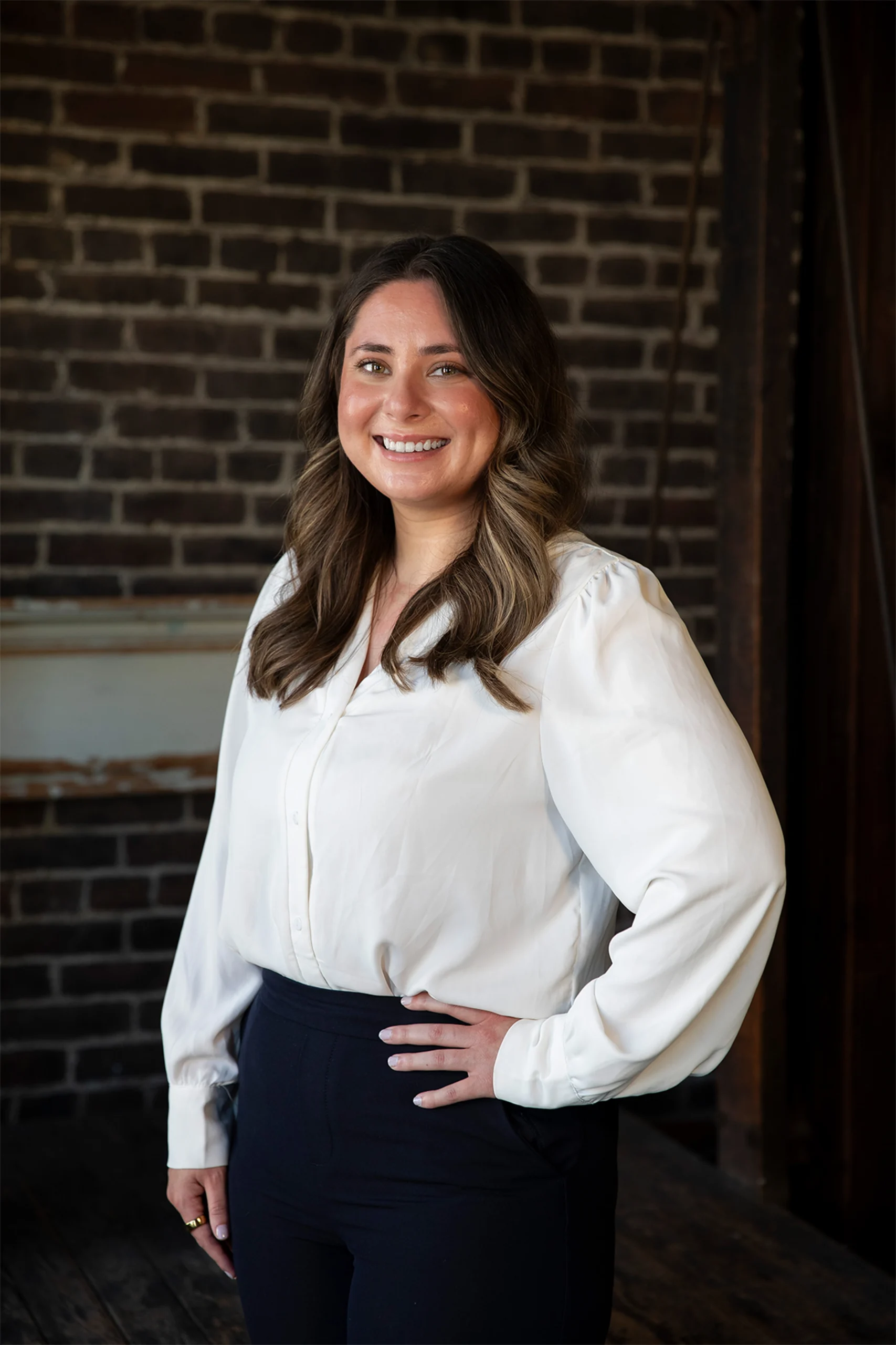 Professional headshot of a smiling woman in a white blouse against an exposed brick wall