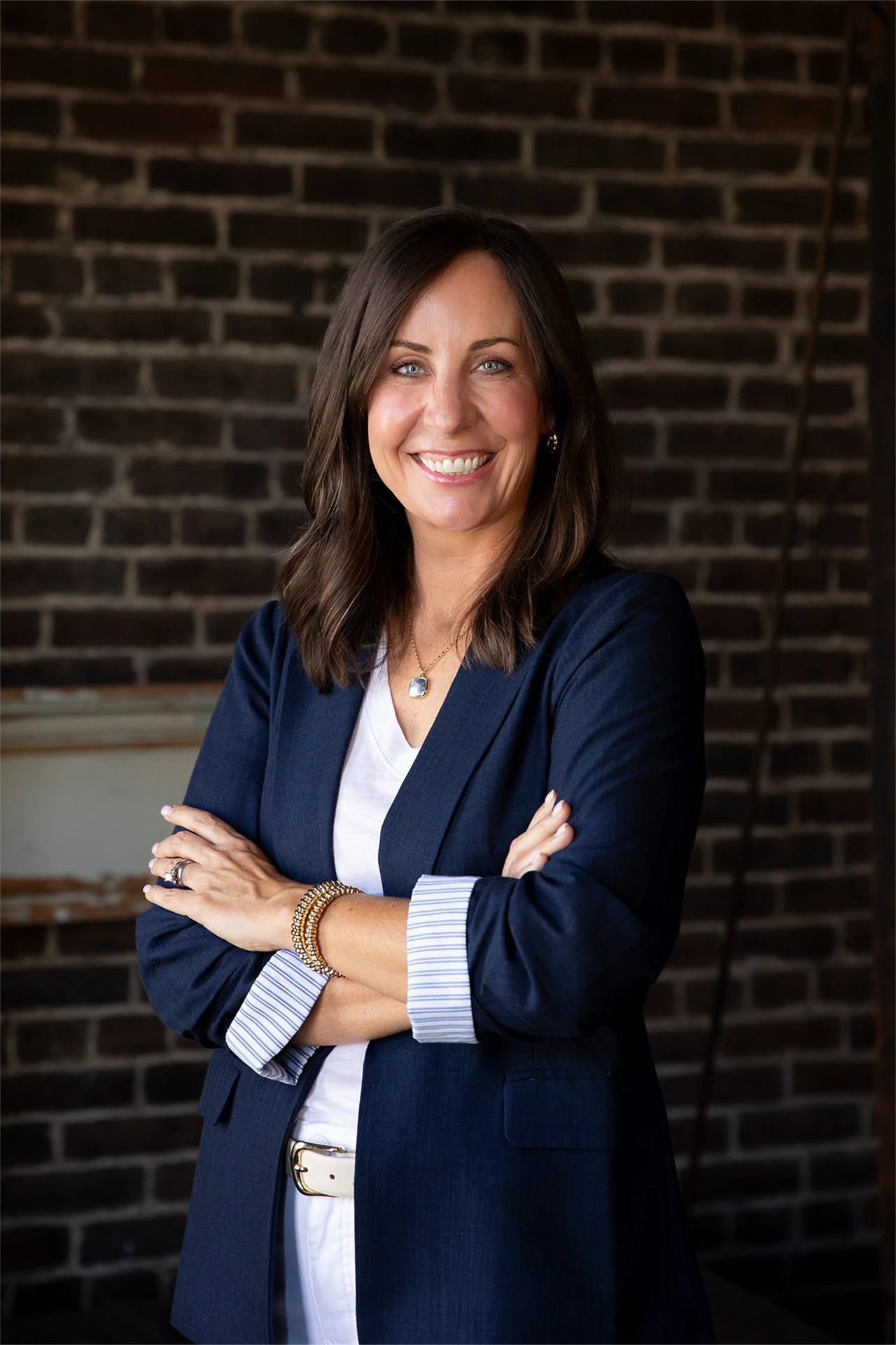 Professional headshot of a smiling woman in a navy blazer against an exposed brick wall