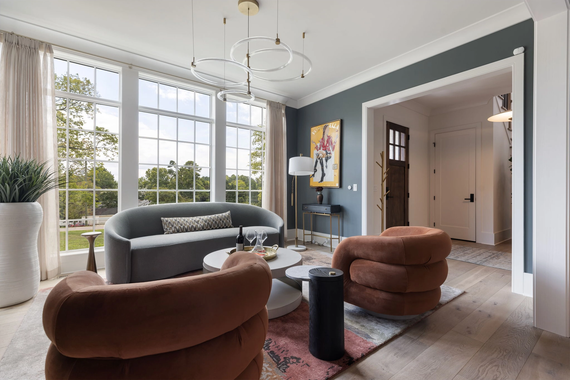 Living room with tall windows, curved gray sofa, brown velvet chairs, and a gold ring chandelier