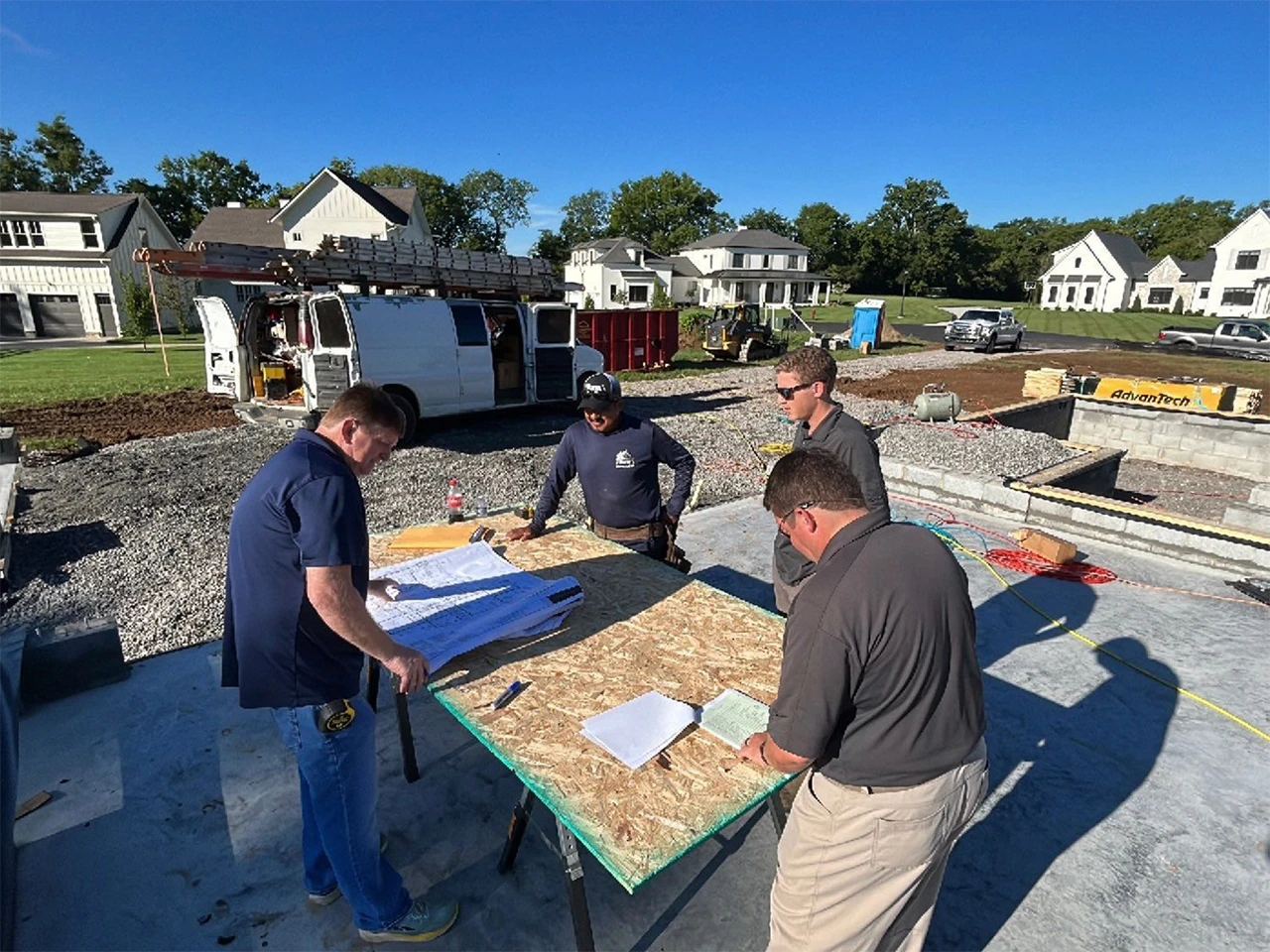 Four workers reviewing plans on a table at a job site with homes in the background