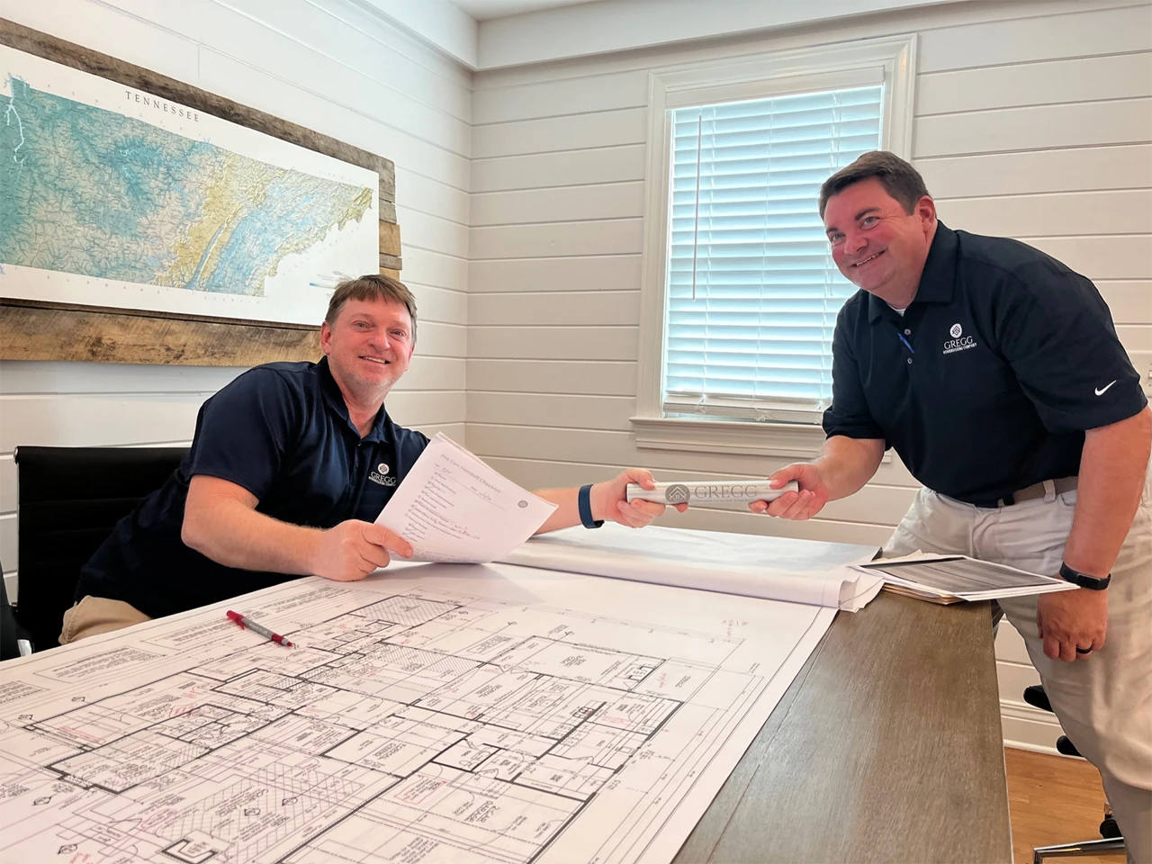 Two men smiling at a desk while reviewing blueprints, with a framed state map on the wall