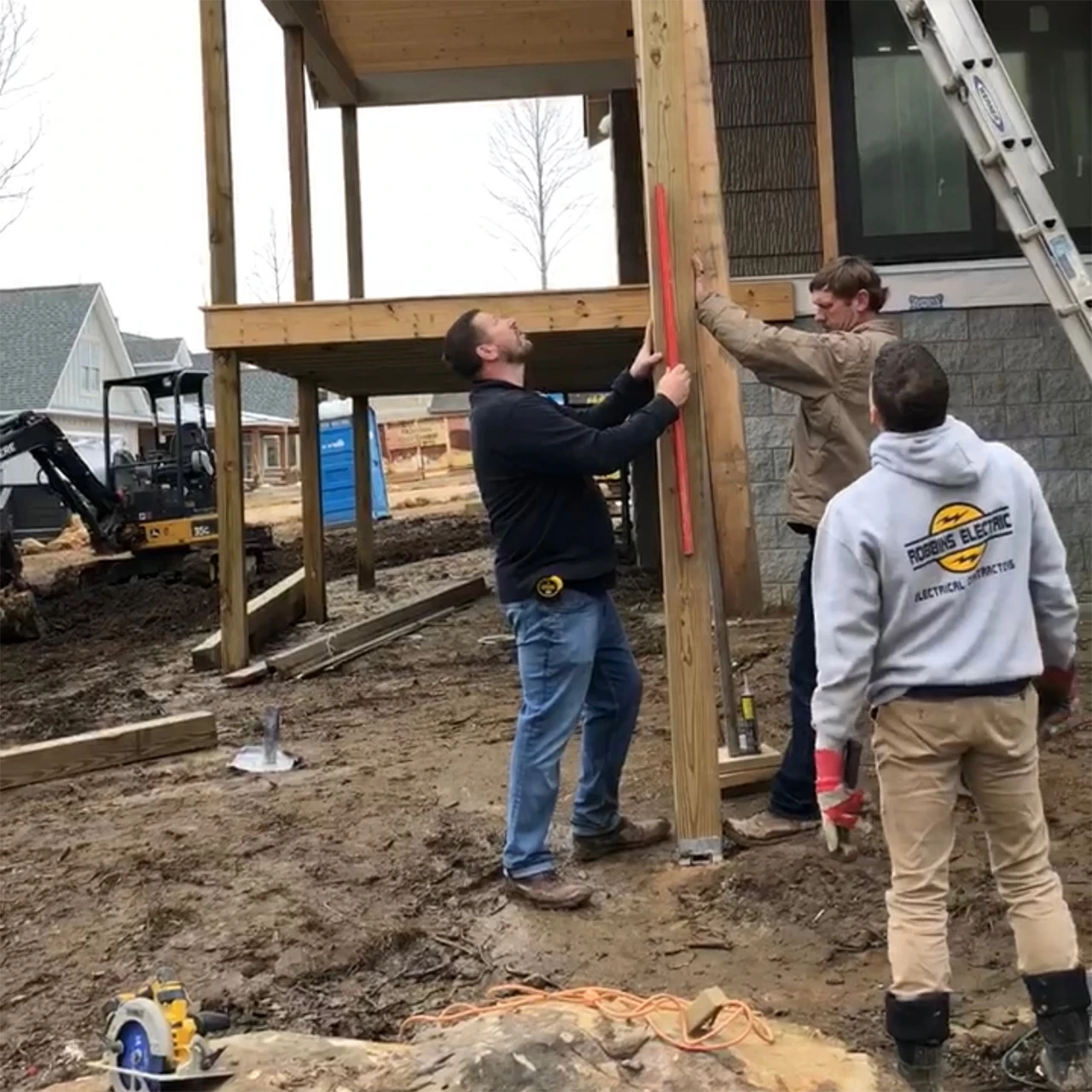 Construction crew installing a wood porch post, one worker holding a level, at a job site