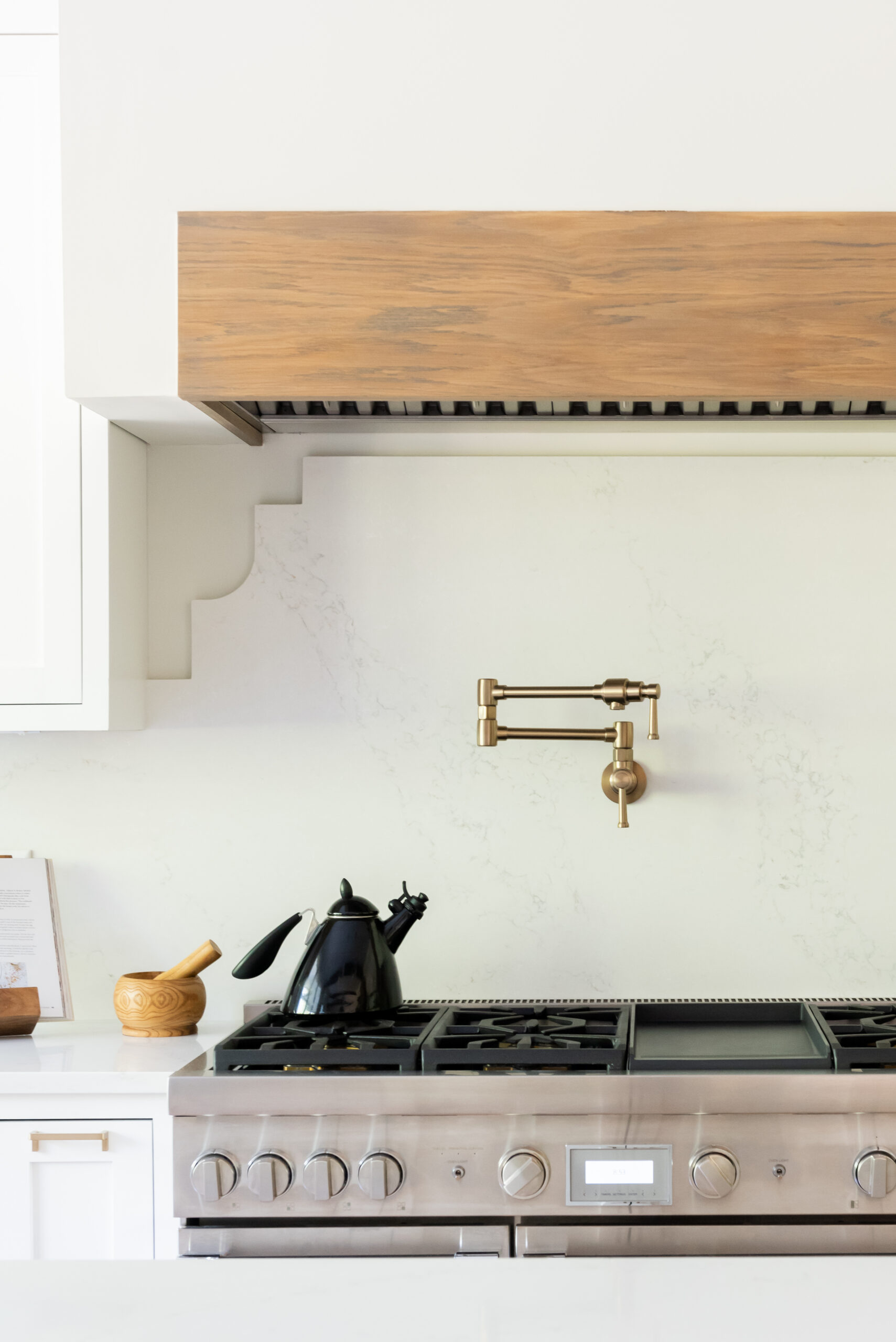 Kitchen range with white backplash and black pot-filler with hood with light stained natural wood header