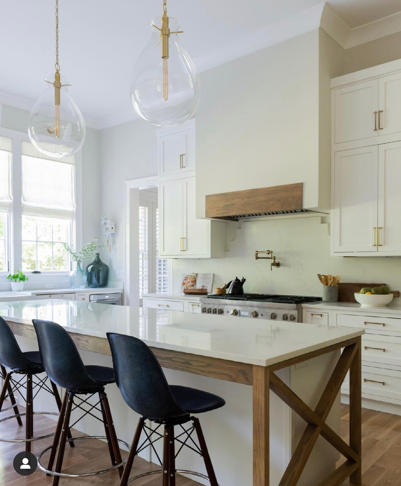 Kitchen range with white backplash and black pot-filler with hood with light stained natural wood header and windows over the sink and clear glass pendant lights