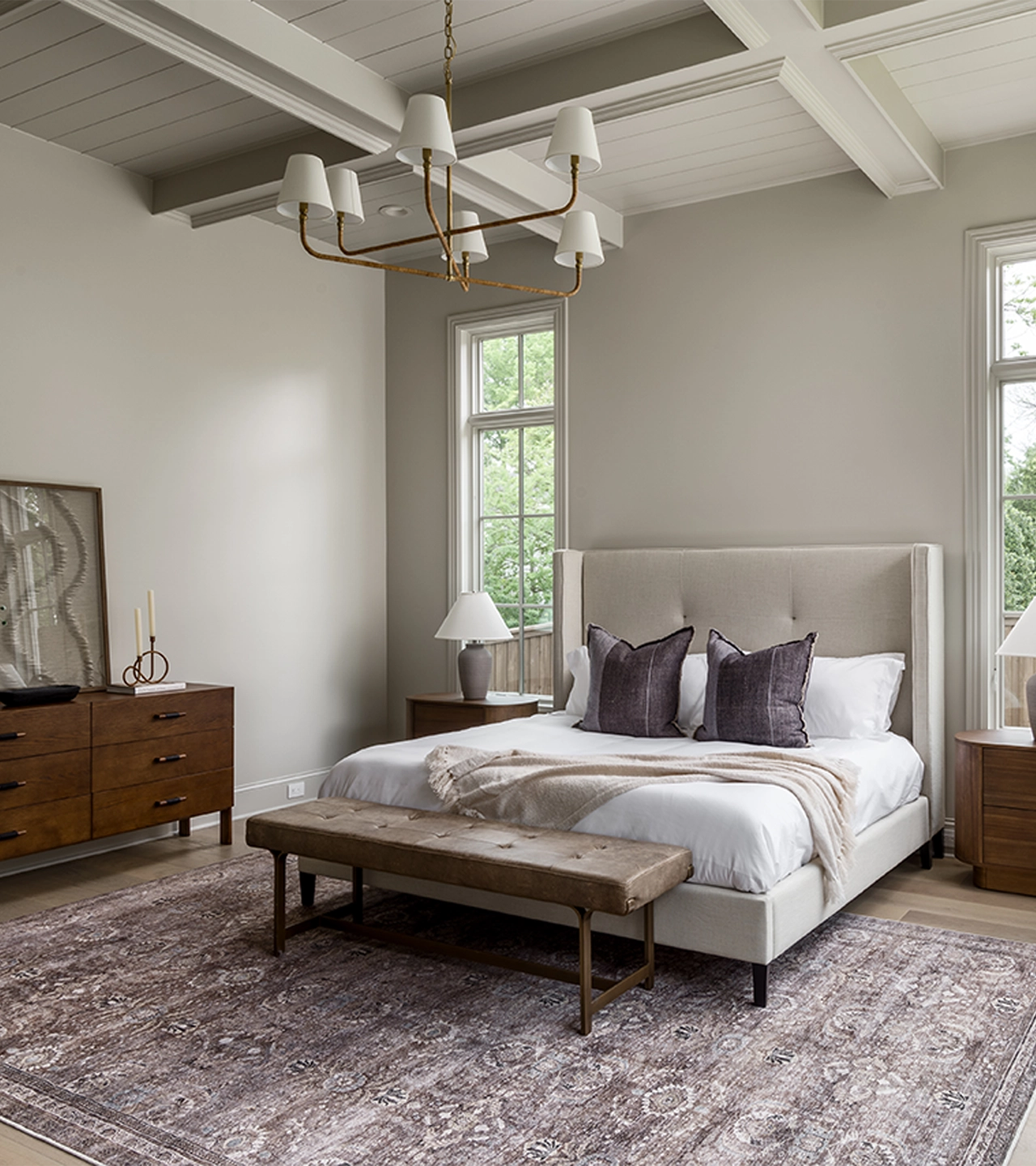 Bedroom with light gray walls and ceiling beams, gray headboard, and a chandelier and rug