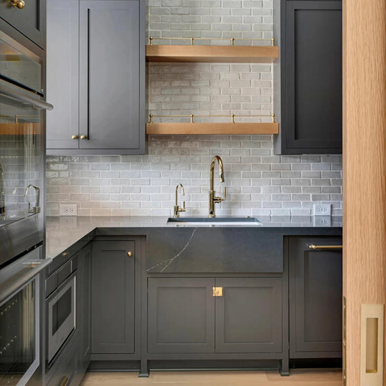 Charcoal gray kitchen with light brick backsplash, brass gooseneck faucet, and floating wood shelves