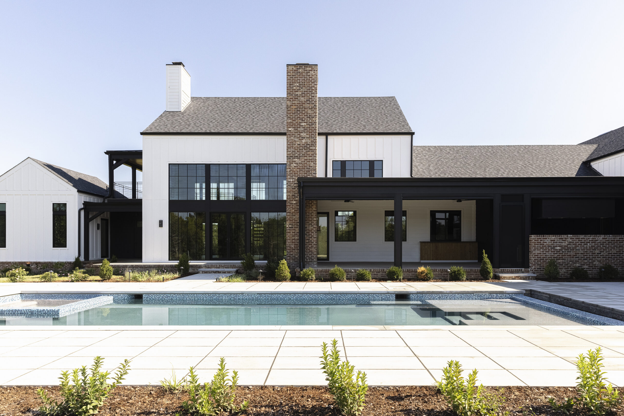 Two story modern farmhouse with white siding and brick with black metal porch railings and large windows and a large rectangular pool