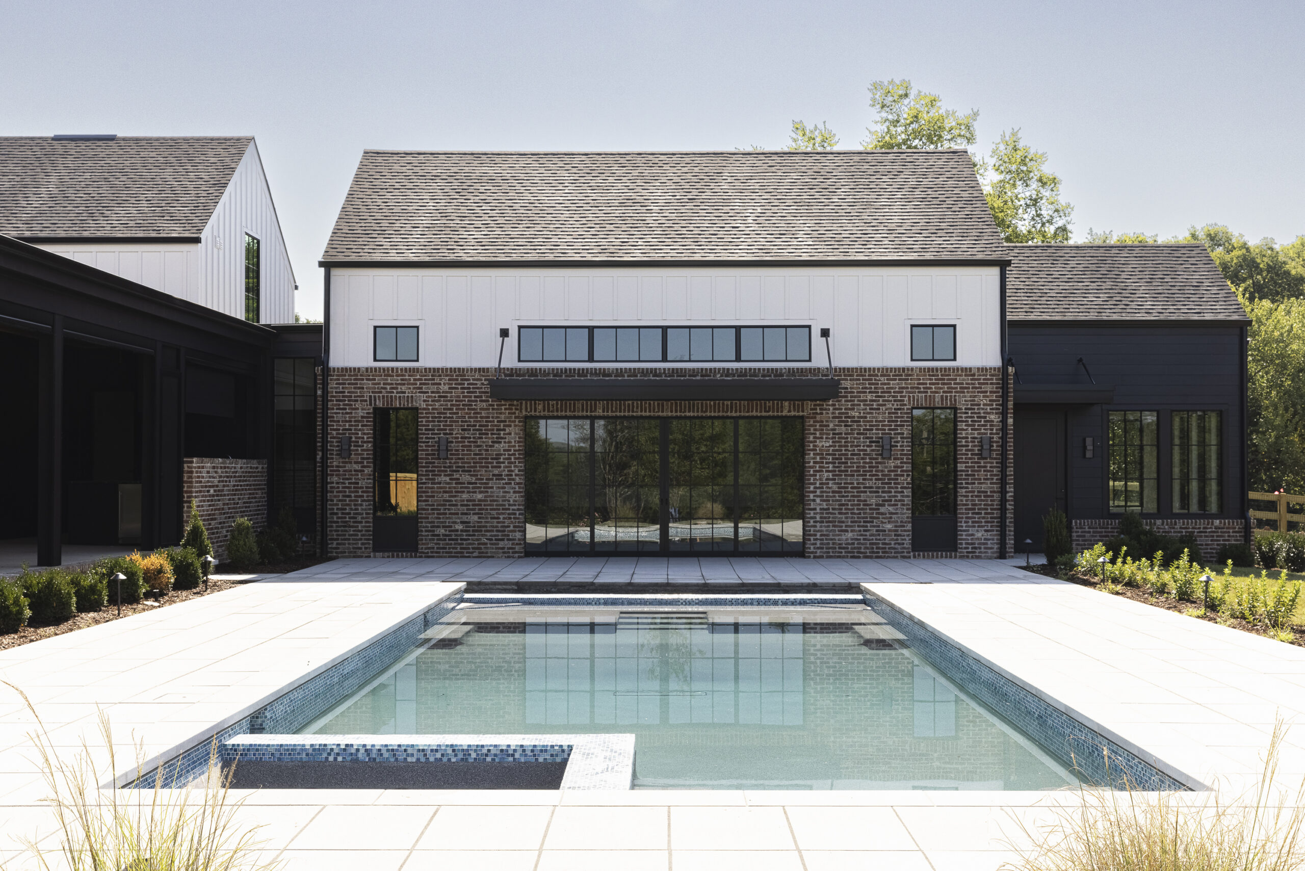 Two story modern farmhouse with white siding and brick with black metal porch railings and large windows and a large rectangular pool