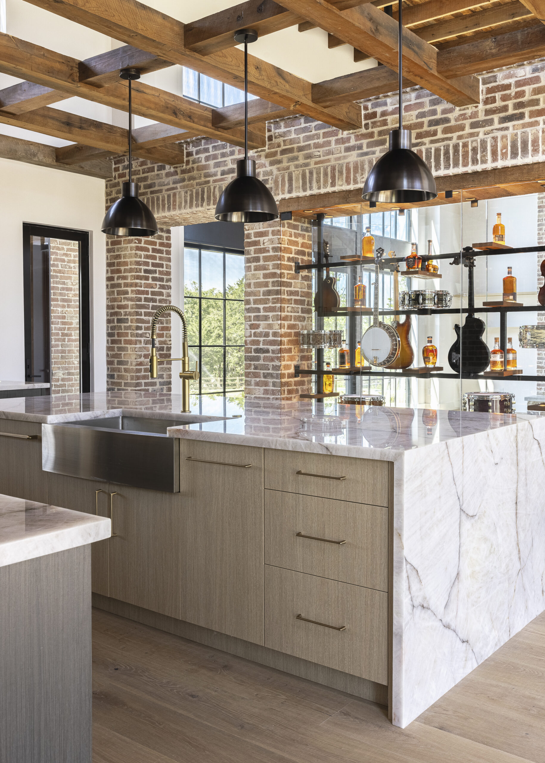 Side view of kitchen with windows on either side of light natural wood stained refrigerator door with oversized brushed gold door and drawer pulls with black pendant lights over a white island with a waterfall edge and wood stained beams overhead