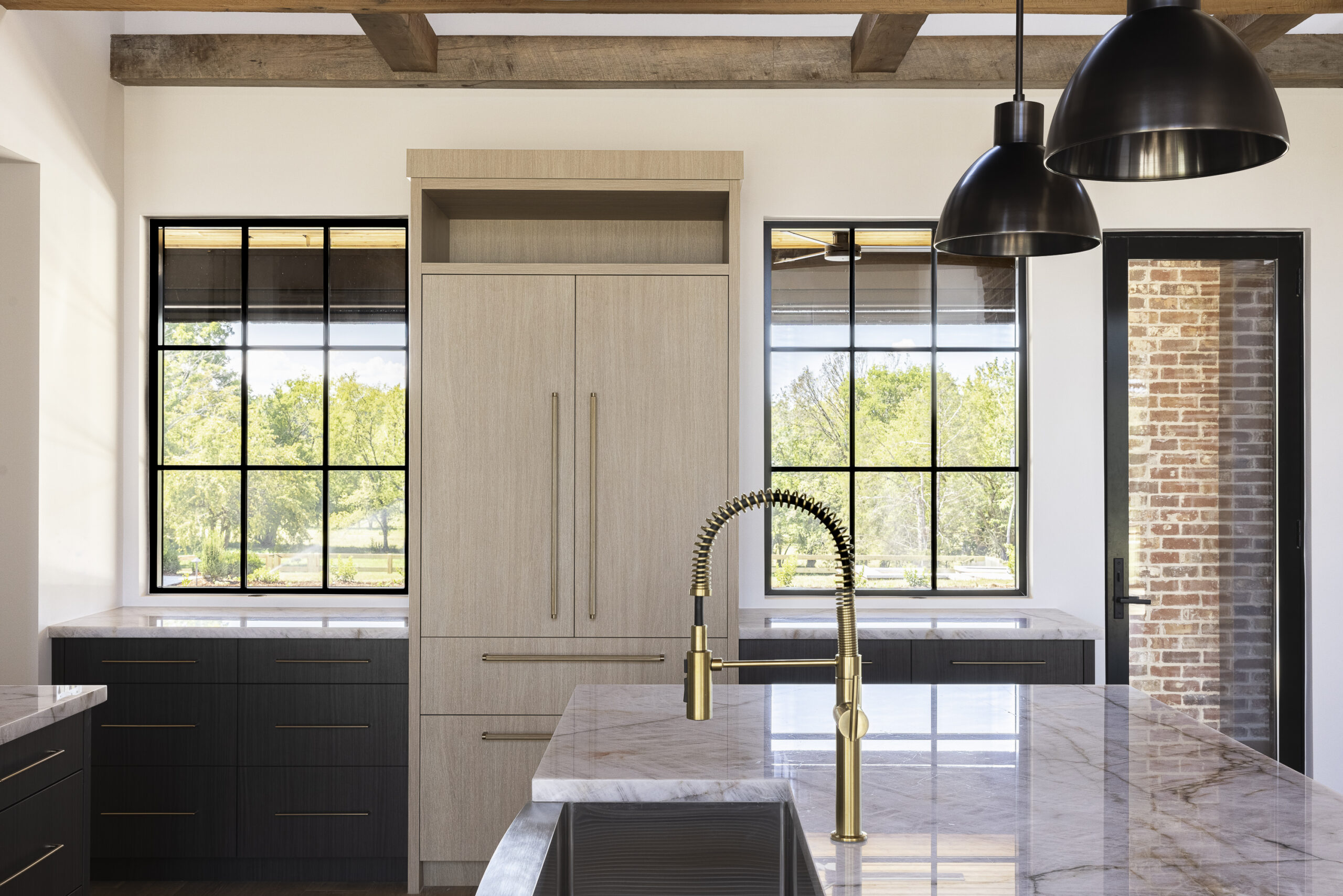 Side view of kitchen with windows on either side of light natural wood stained refrigerator door with oversized brushed gold door and drawer pulls with black pendant lights over a white island with a waterfall edge and wood stained beams overhead
