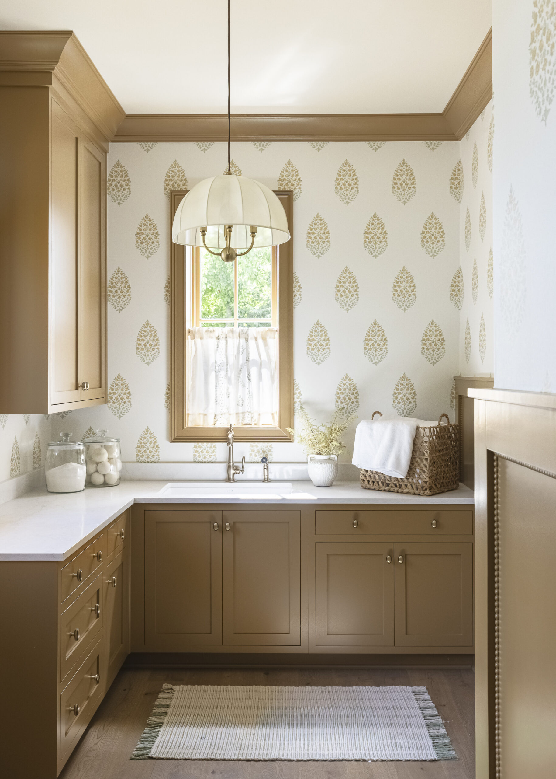 Warm orche cabinetry in laundry room with white stamped wall paper and window with trim painted the same as the cabinetry and white counterops with chandlier with white umbrella shade