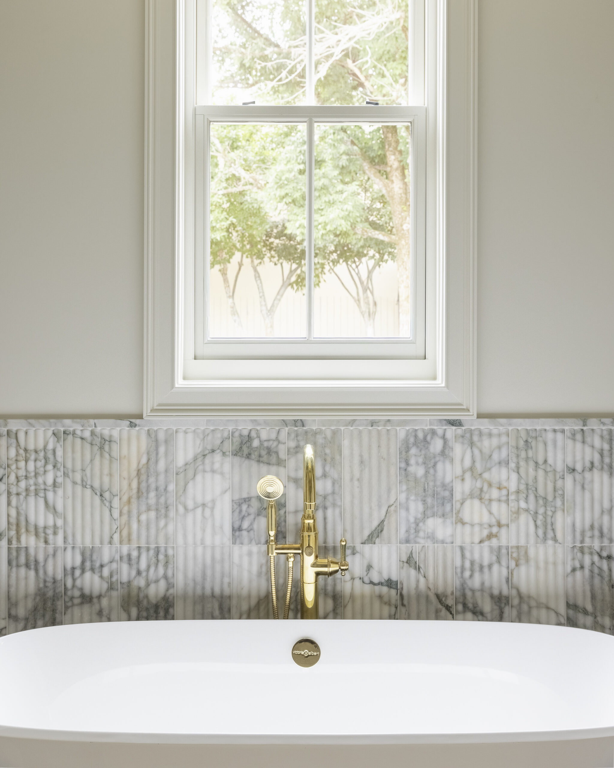Fluted marble framing and halfwall behind white tub with gray tile and light gray cabinetry