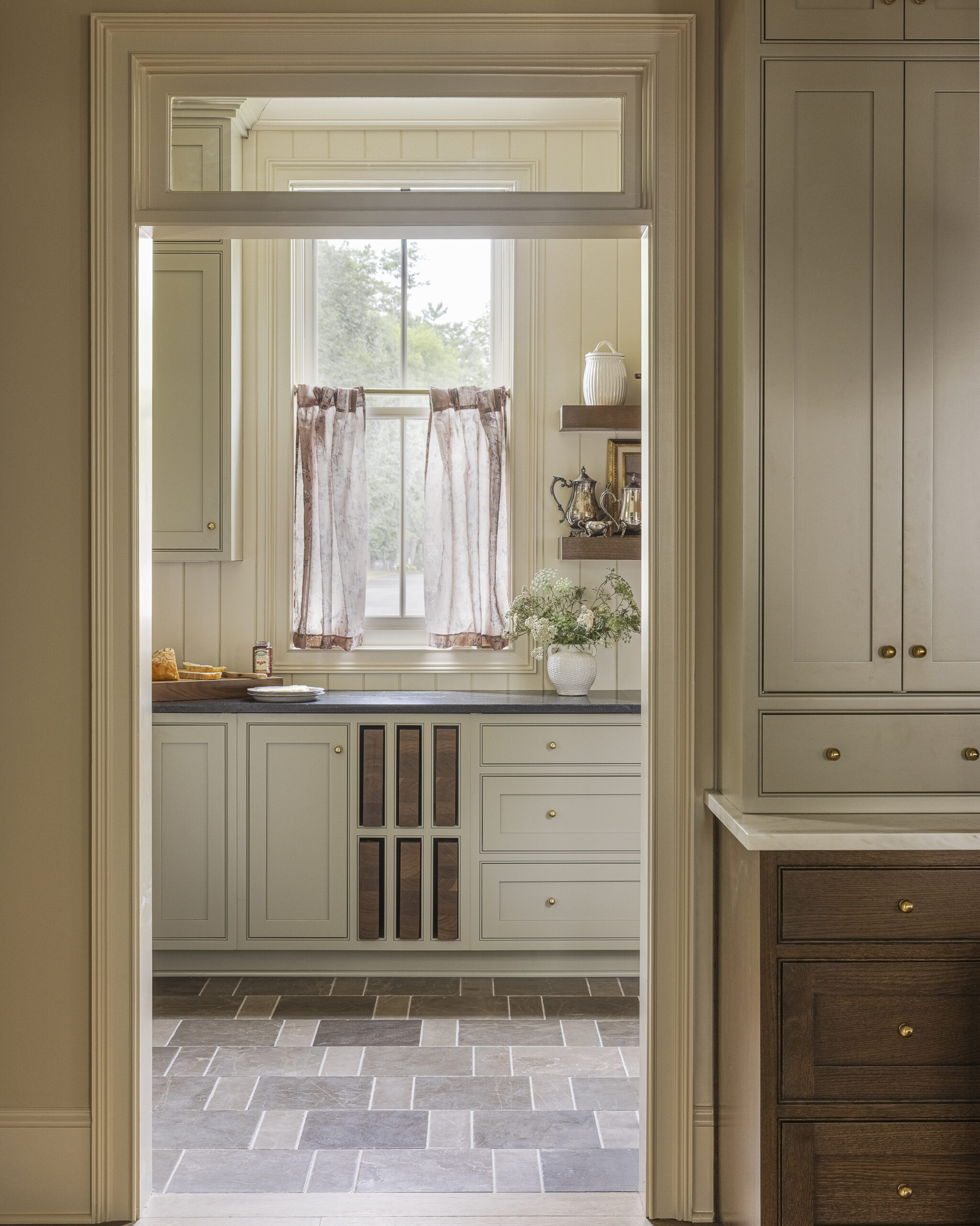 Scullery with sage cabinets, black soapstone cabinets and v-groove cream wall paneling and window with decorative brass wall sconces