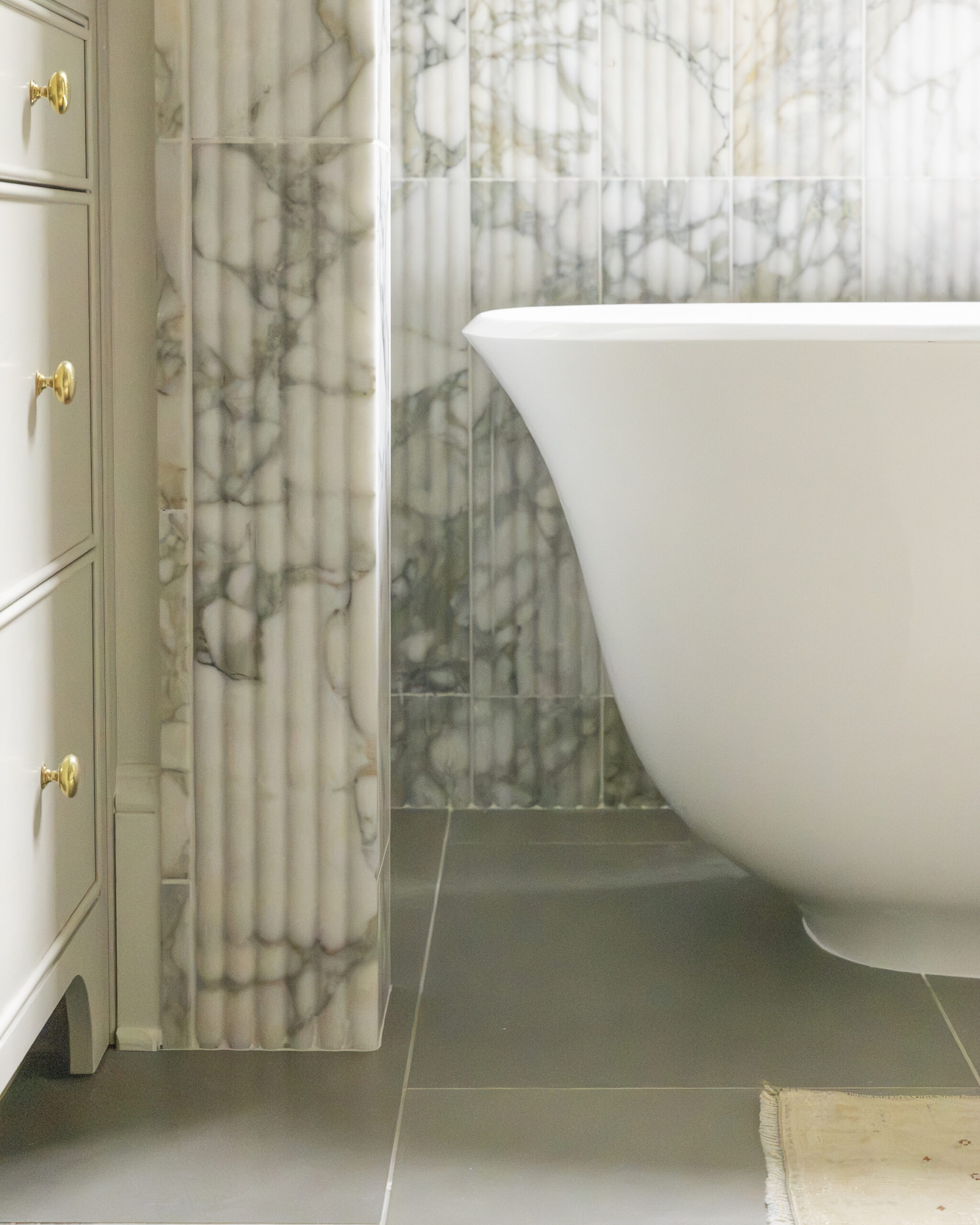 Fluted marble framing and halfwall behind white tub with gray tile and light gray cabinetry