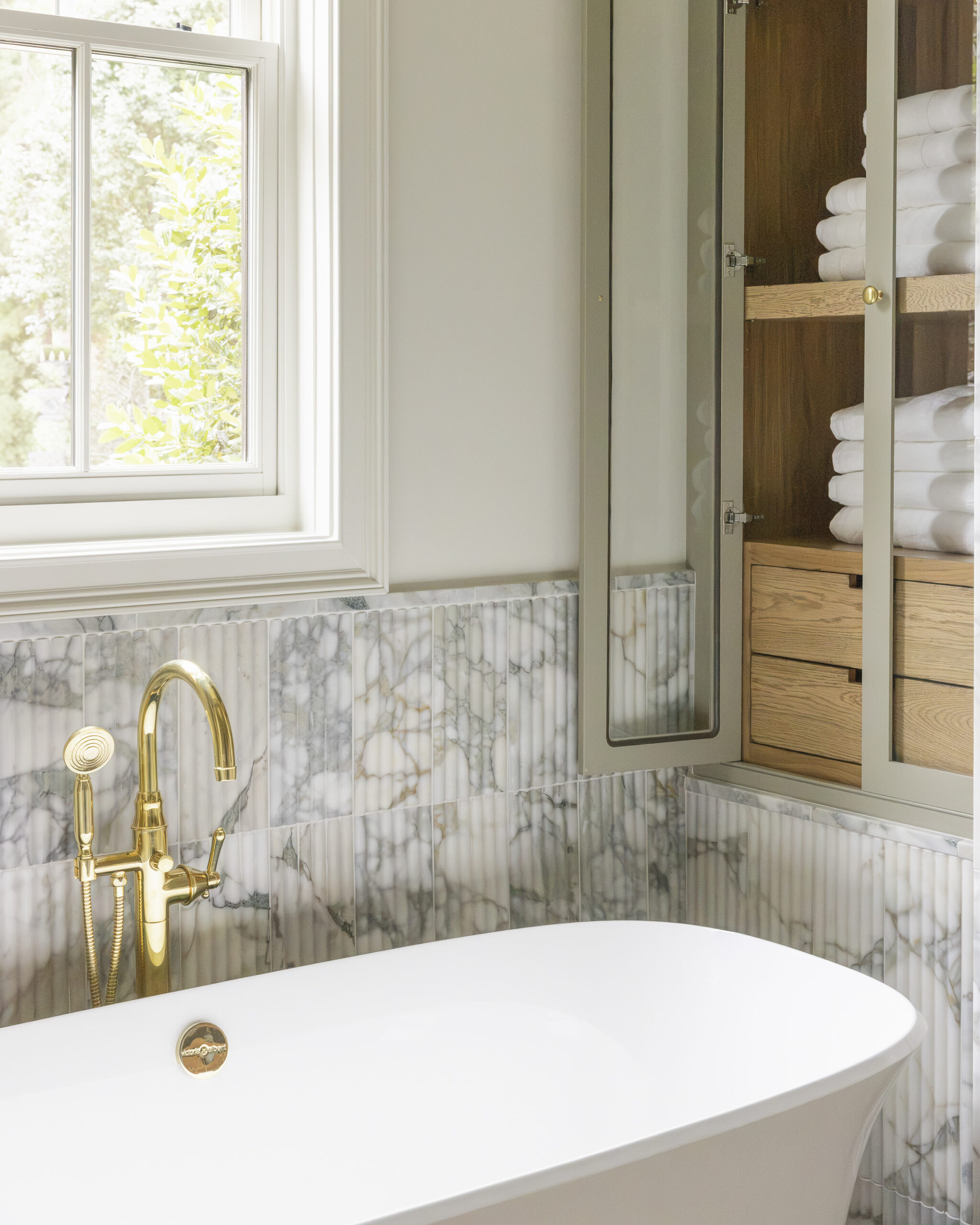 Fluted marble framing and halfwall behind white tub with gray tile and light gray cabinetry