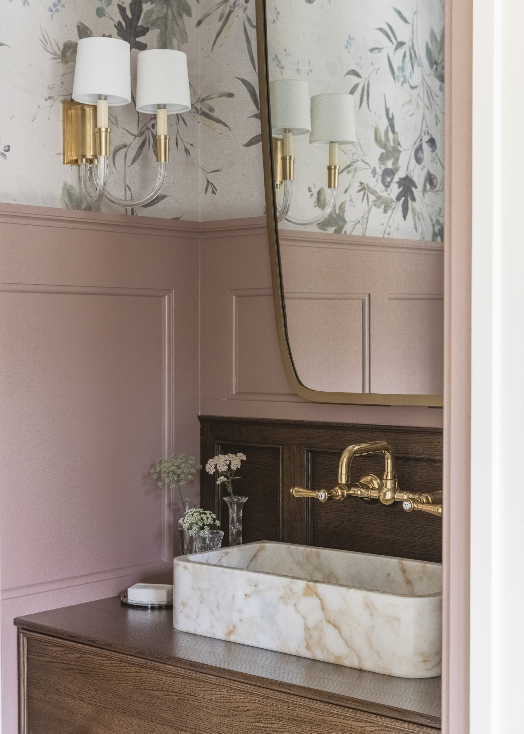 Bathroom with wood stained vanity with brushed gold fixtures a mirror and sconces, mauve pink paneling and floral wallpaper