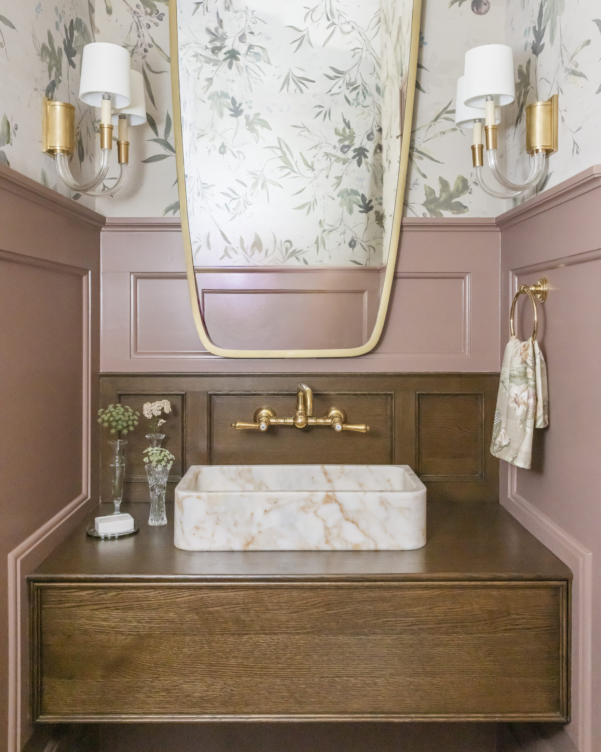 Bathroom with wood stained vanity with brushed gold fixtures a mirror and sconces, mauve pink paneling and floral wallpaper