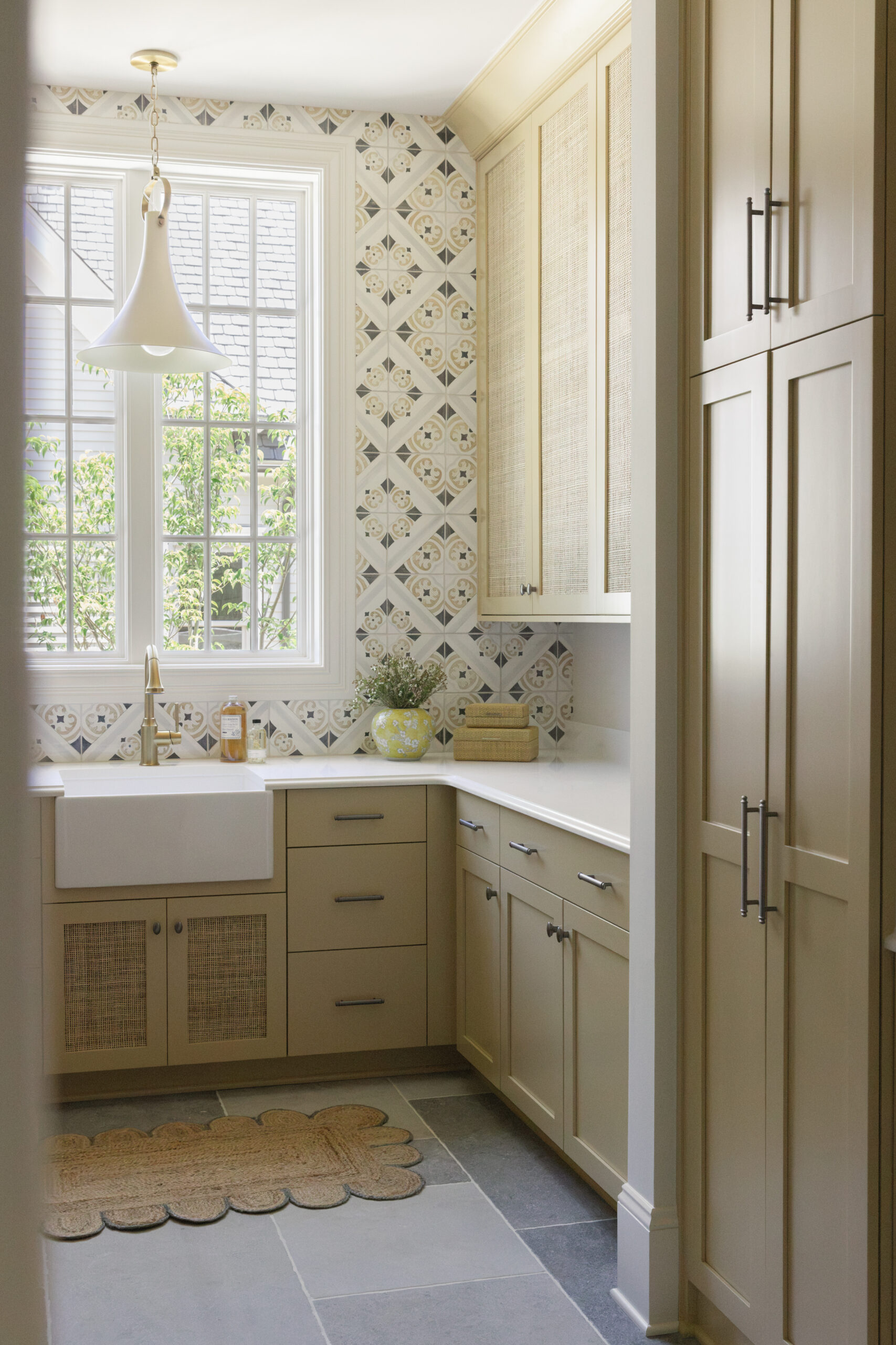 Warm ochre cabinetry in laundry room with patterned tile backsplash and window and white counterops small utility sink