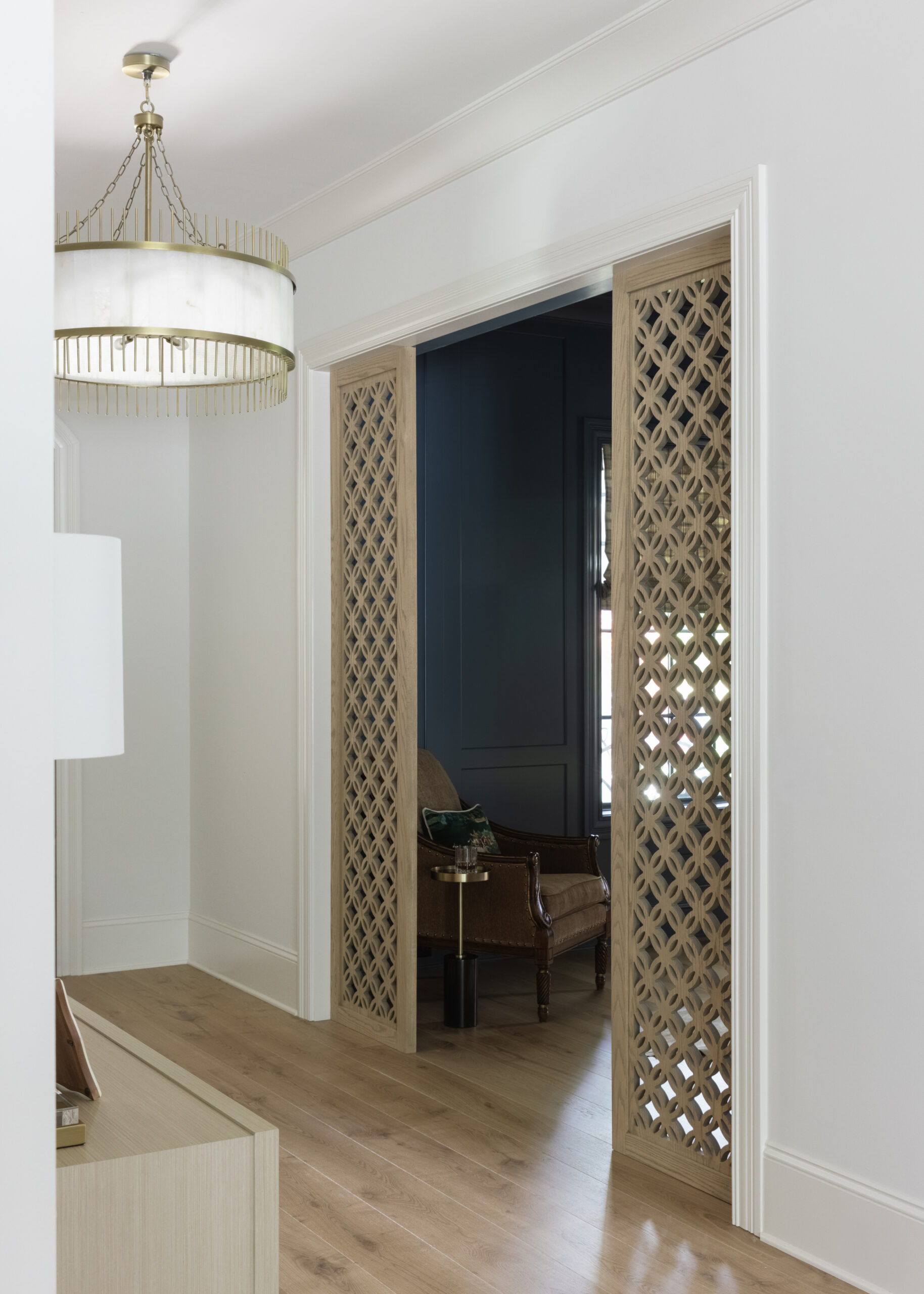 Entryway with white walls and round glass chandelier with wood carved panels framing opening into home office