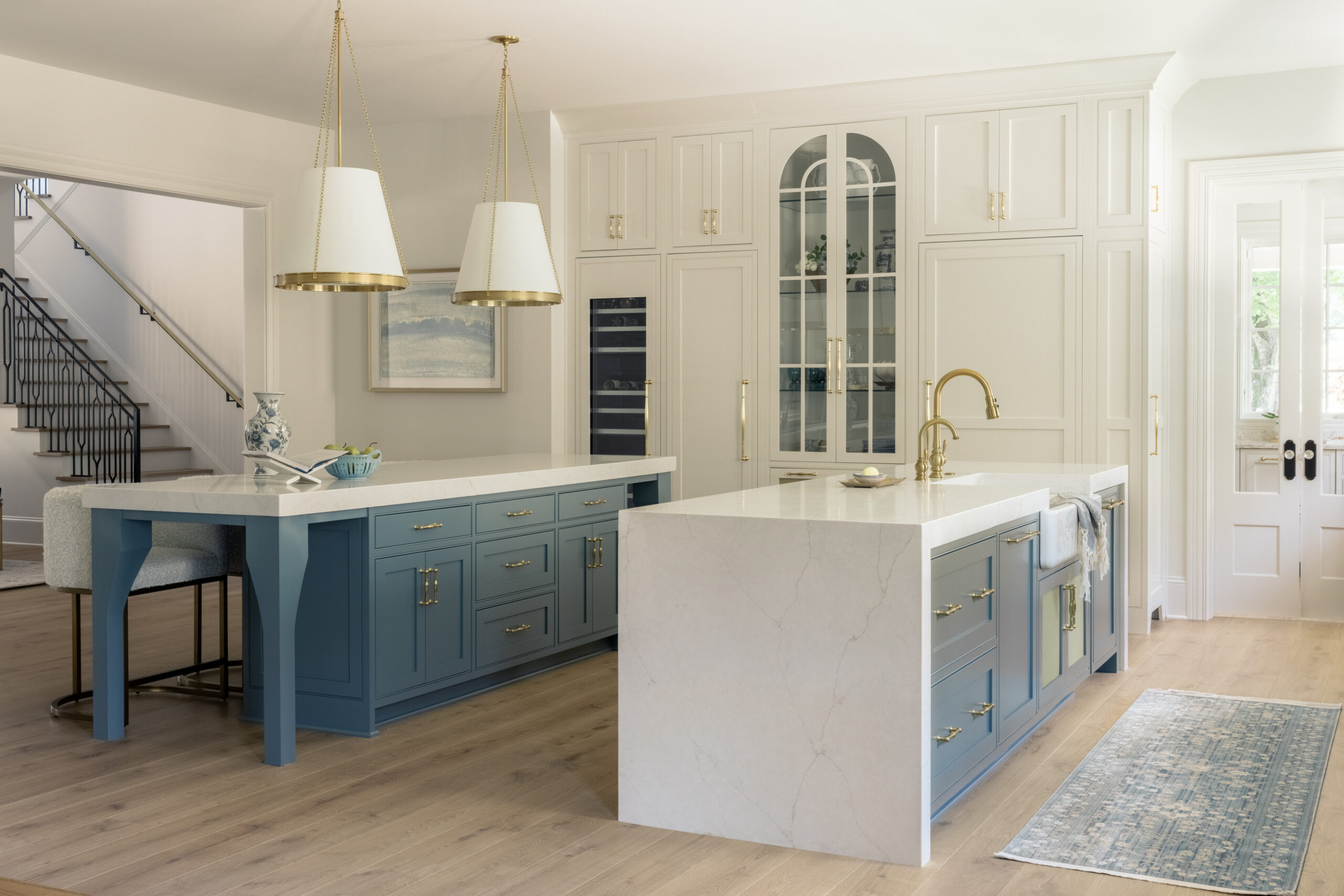 Side view of kitchen with double blue kitchen island cabinets and white countertops with white shaded pendants and glass front pantry cabinet