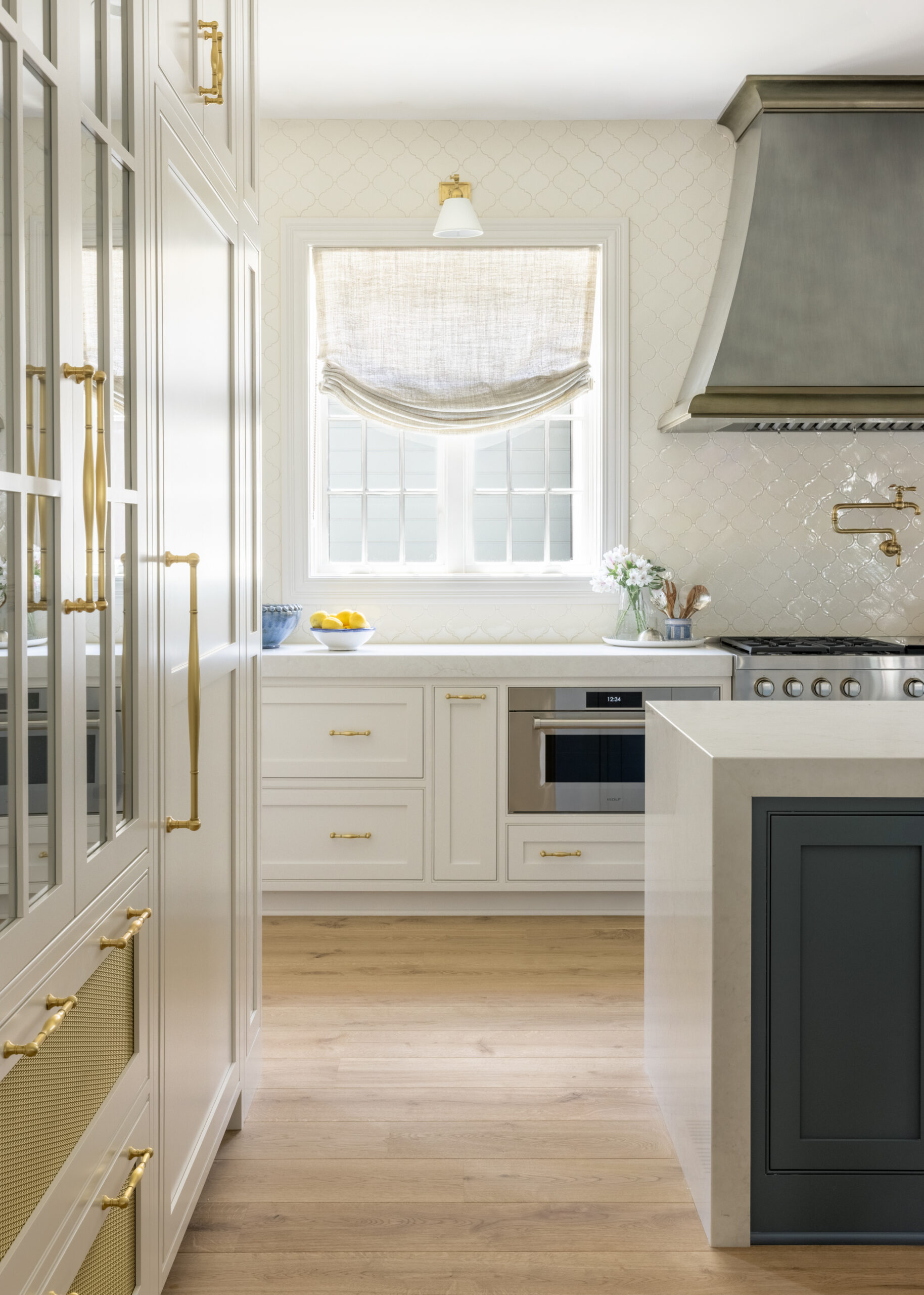 Kitchen view of white cabinets with a glass front pantry cabinet and window beside silver metallic range hood