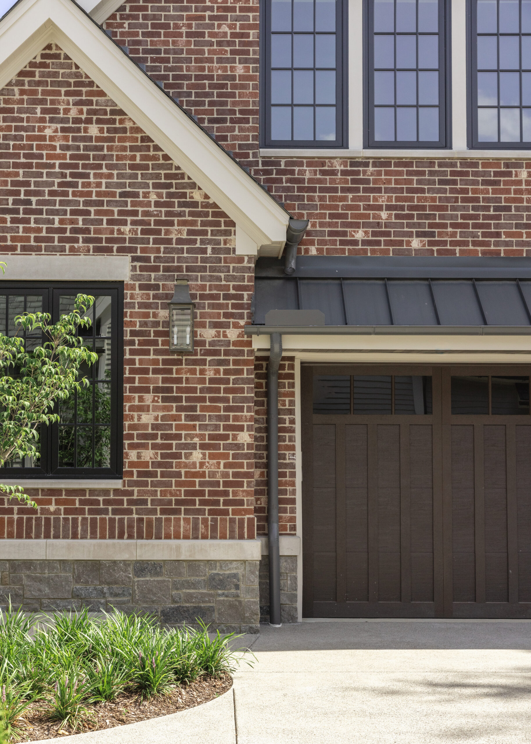 Side of brick home with metal roof overhang over wood stained garage doors and black grid windows