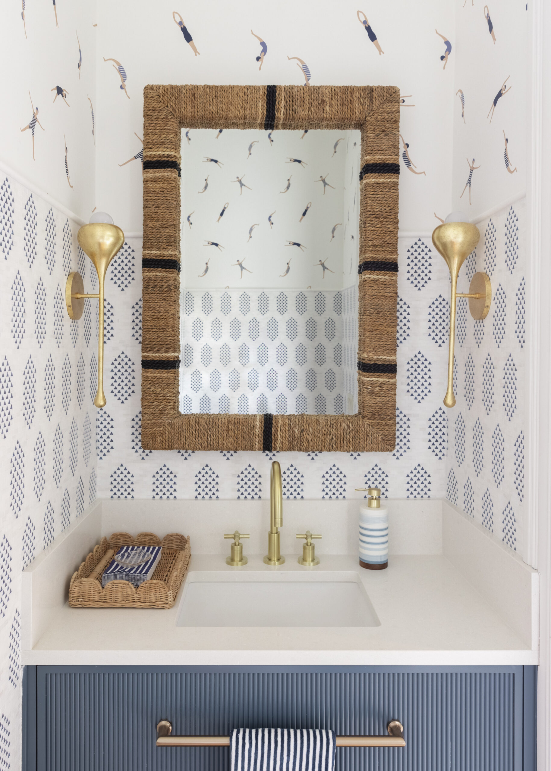 Blue vanity with white countertops and blue and white patterned tile with swimming themed wallpaper and rope wrapped mirror edge