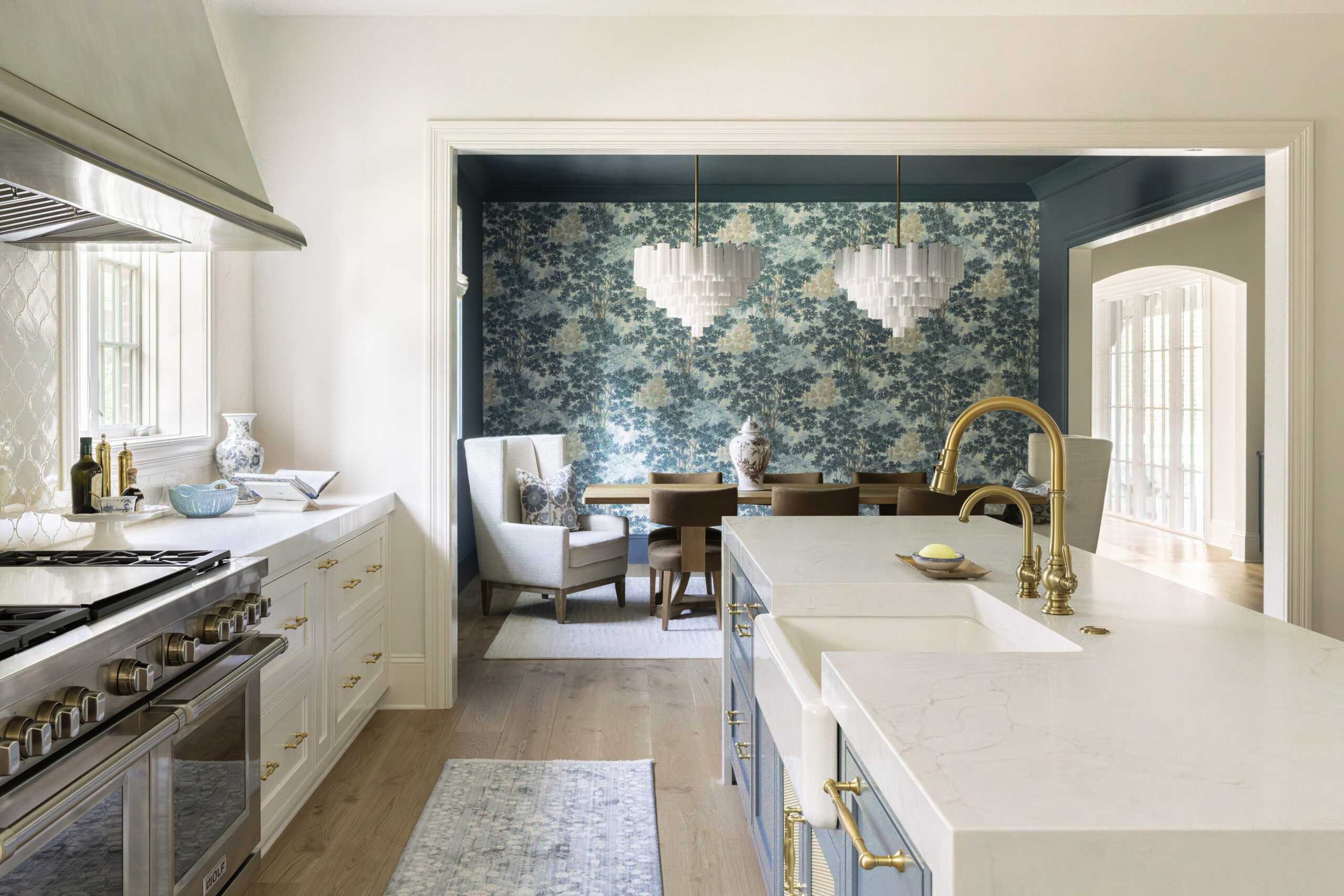 Side view of kitchen with blue kitchen island cabinets and white countertops looking into dining area with blue floral wall paper and twin white glass chandeliers