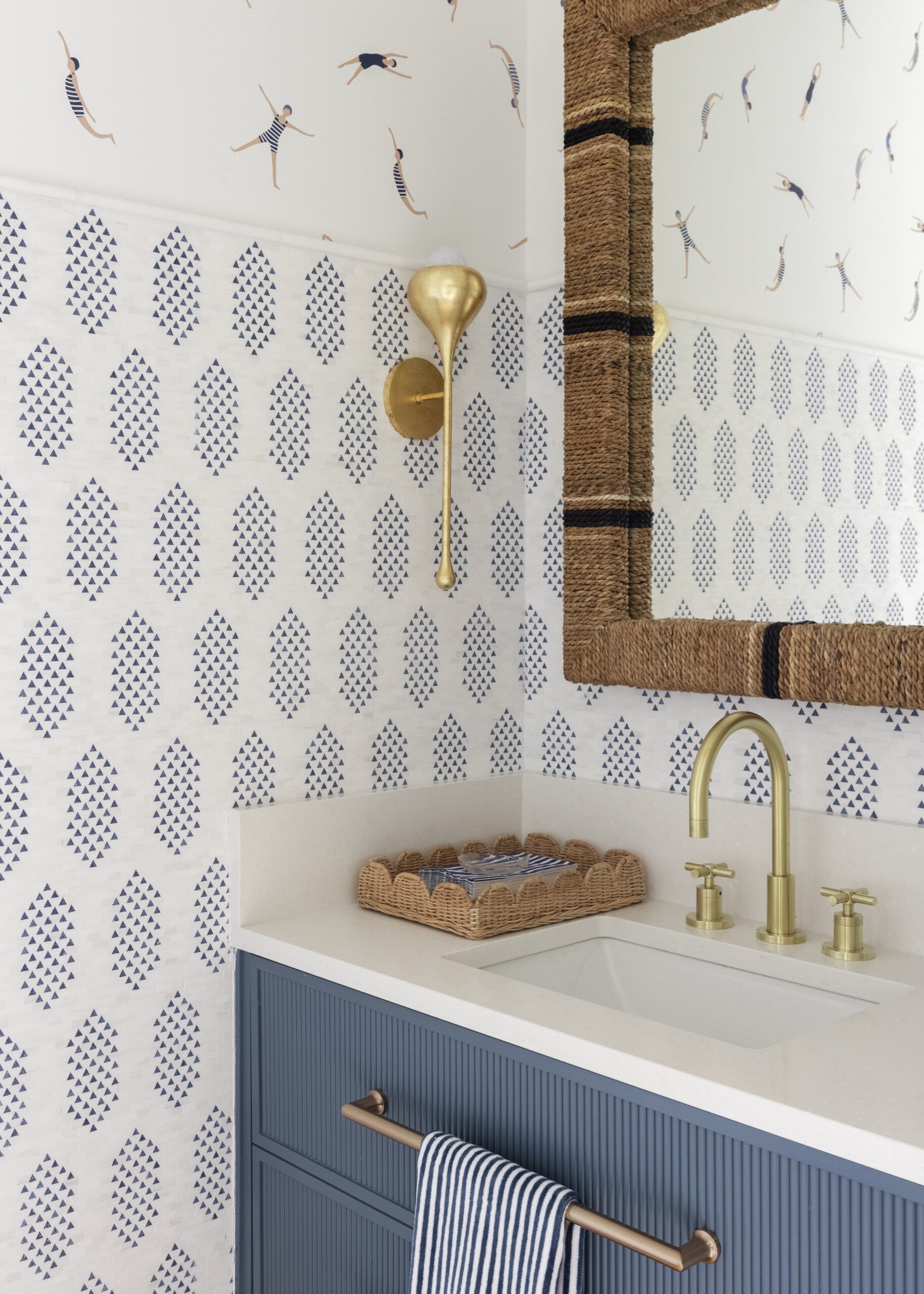 Blue vanity with white countertops and blue and white patterned tile with swimming themed wallpaper and rope wrapped mirror edge