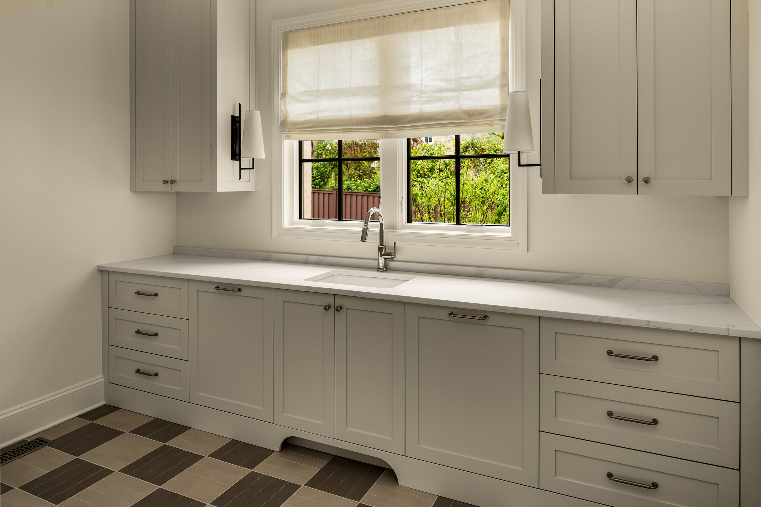 Laundry room with white lower cabinets and white countertops with window above framed with shaded sconces and brown and cream checkered, tile flooring