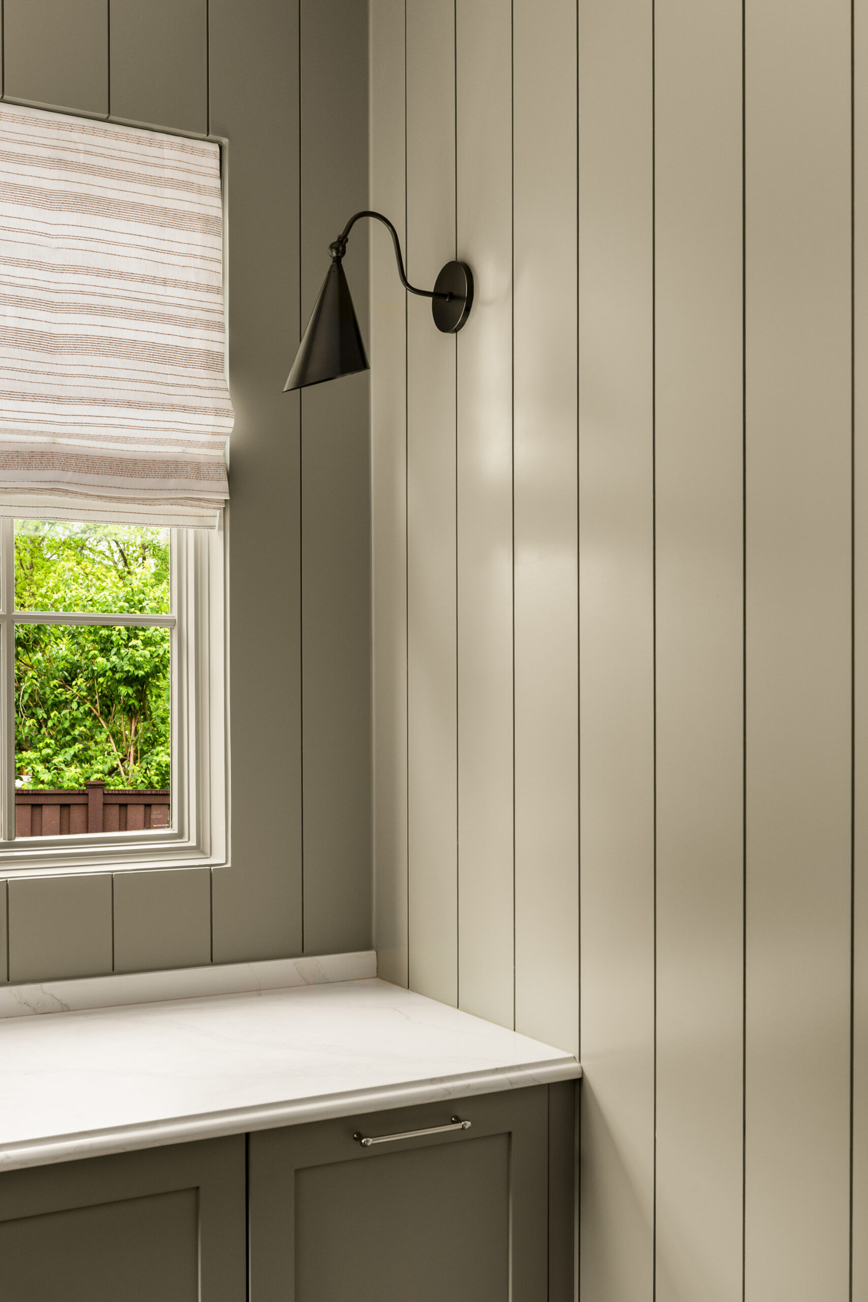 Muted sage green paneled wall with black sconce, light countertops, and window