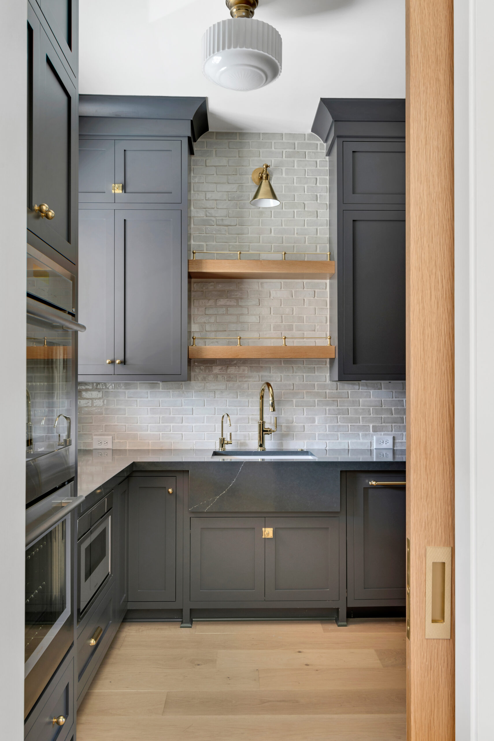 Pantry with navy cabinets and light marble tile backsplash with wood tone open shelving above the utility sink, and stacked double ovens