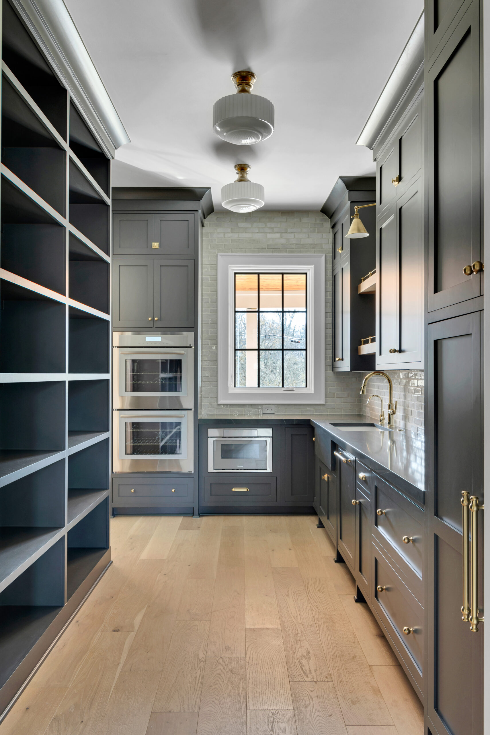 Butler's pantry with navy cabinets, double stacked ovens, floor to ceiling open shelving, utility sink, and light colored countertops and chandelier