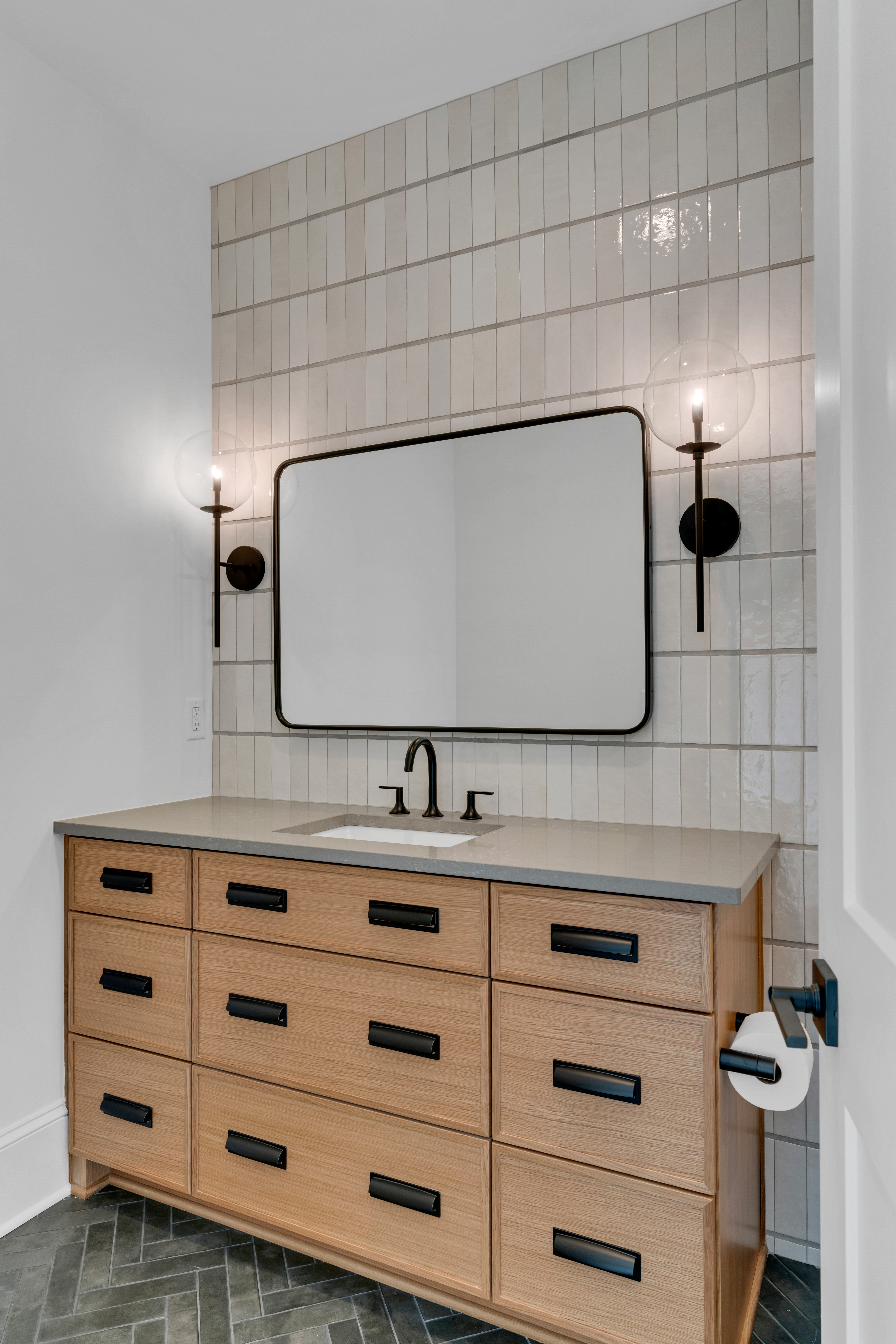 Black tile floor with large vanity and rectangular mirror and tiled wall with sconces