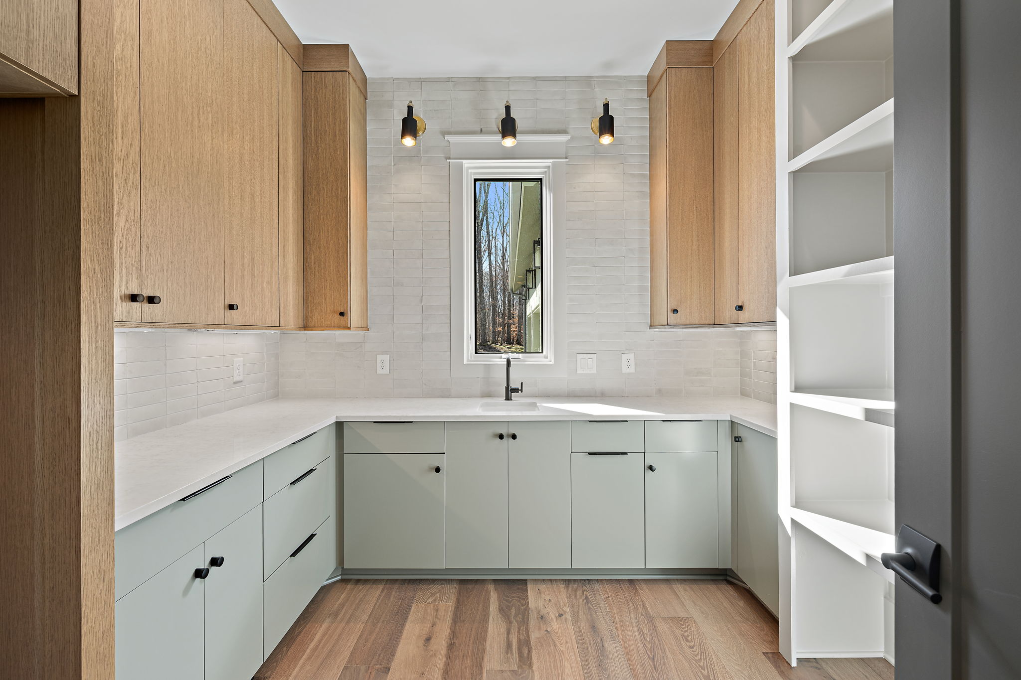 Window with sconce lighting above and marble tile backsplash, with light wood toned upper cabinets and mint base cabinetry